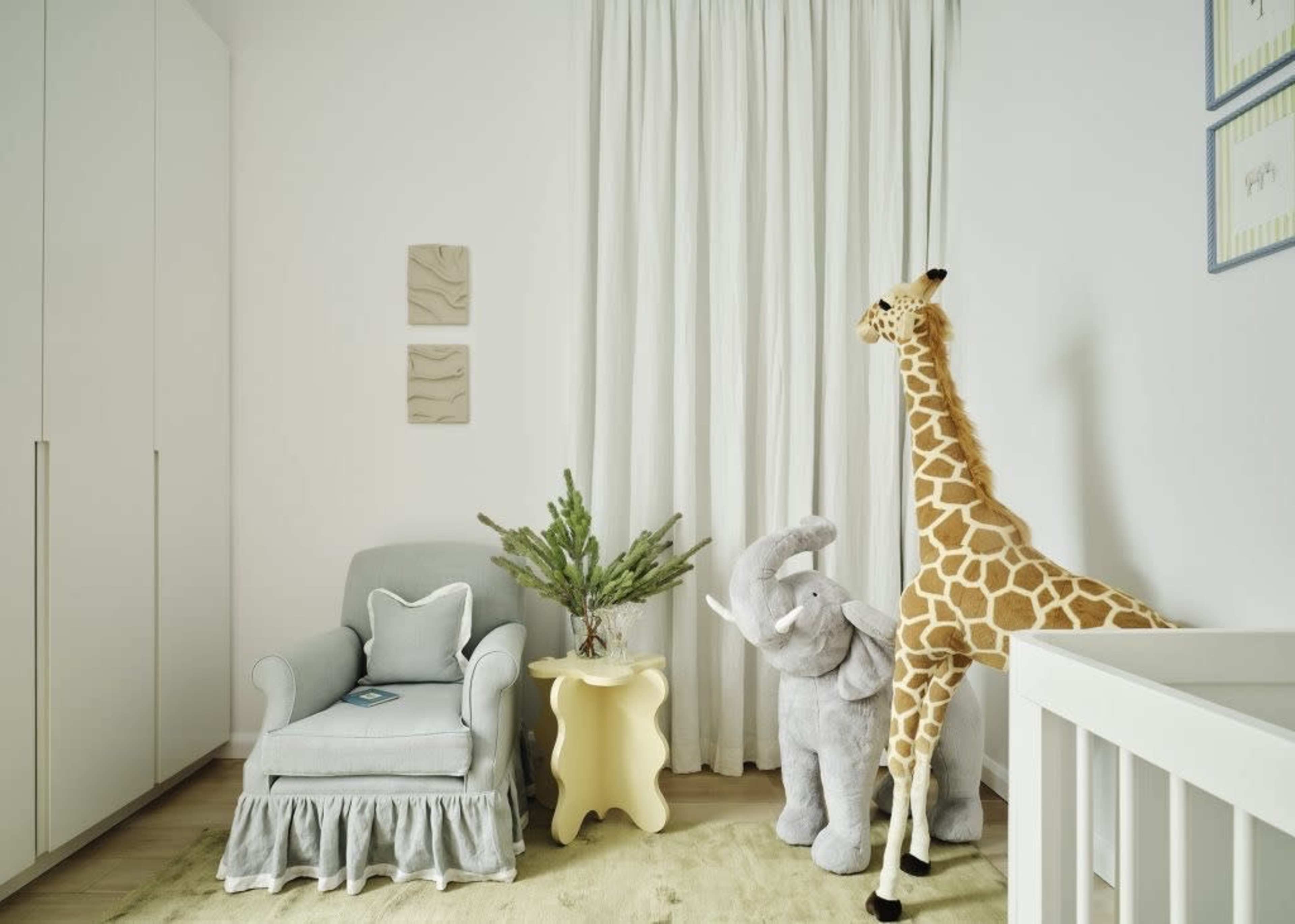 The image shows a nursery with a gray armchair, a white crib, a yellow side table, and large stuffed animals, including a giraffe and an elephant, beside a potted plant.