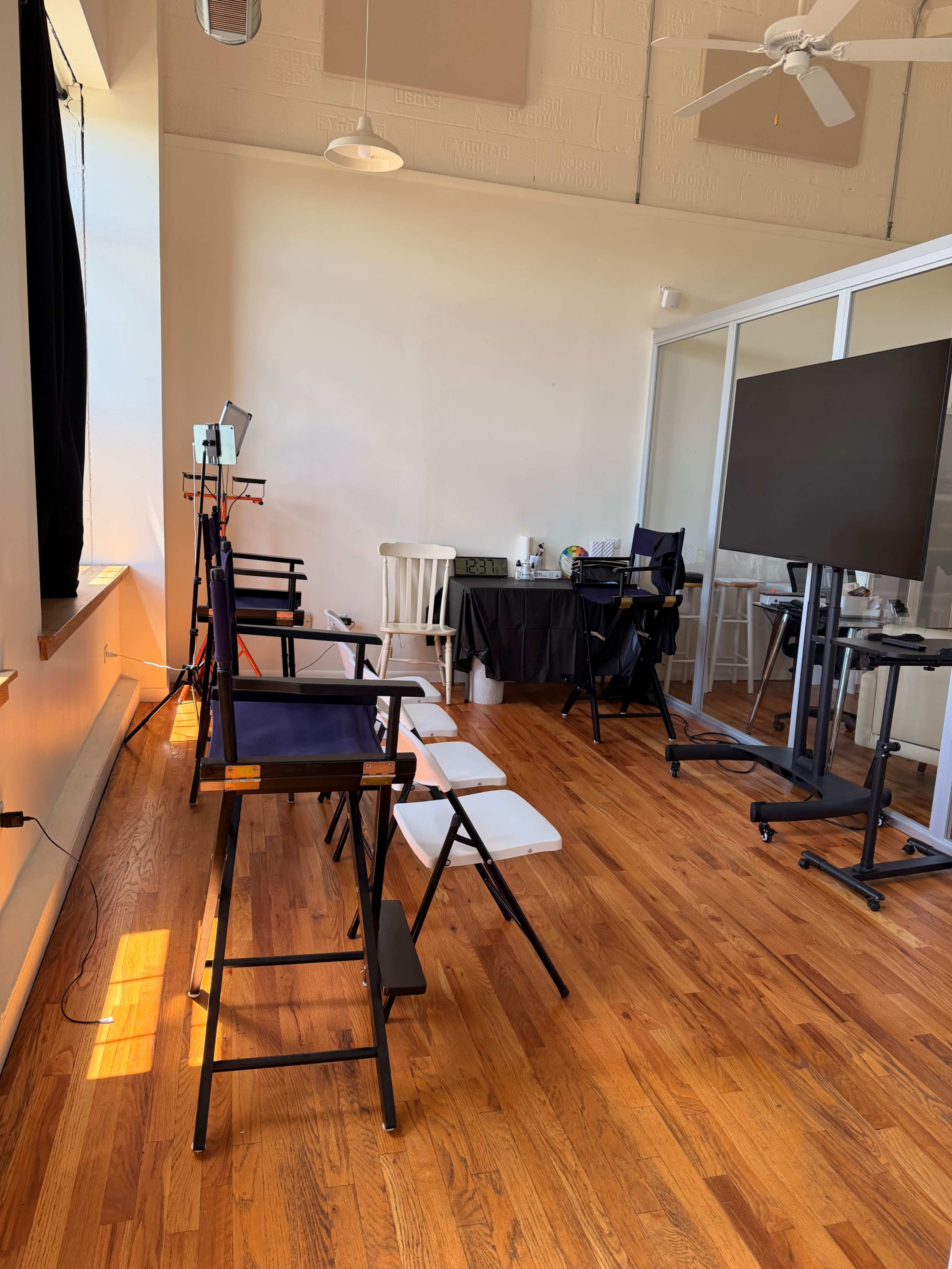 The image shows a well-lit, empty room with wooden floors, featuring a few chairs, a table with equipment, and a large screen on a stand.