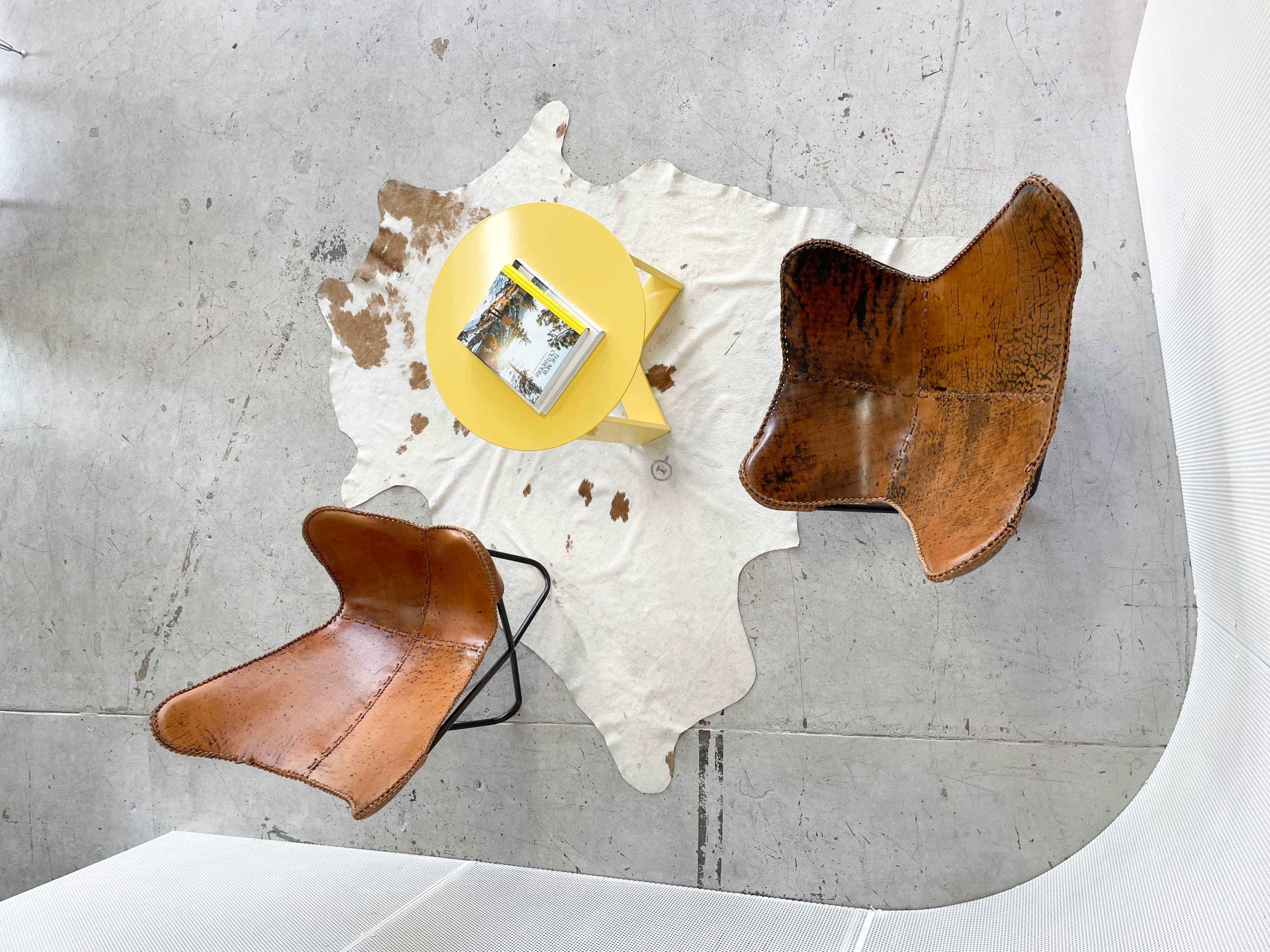 Two brown chairs with a yellow side table are arranged on a white cowhide rug in a minimalist space.