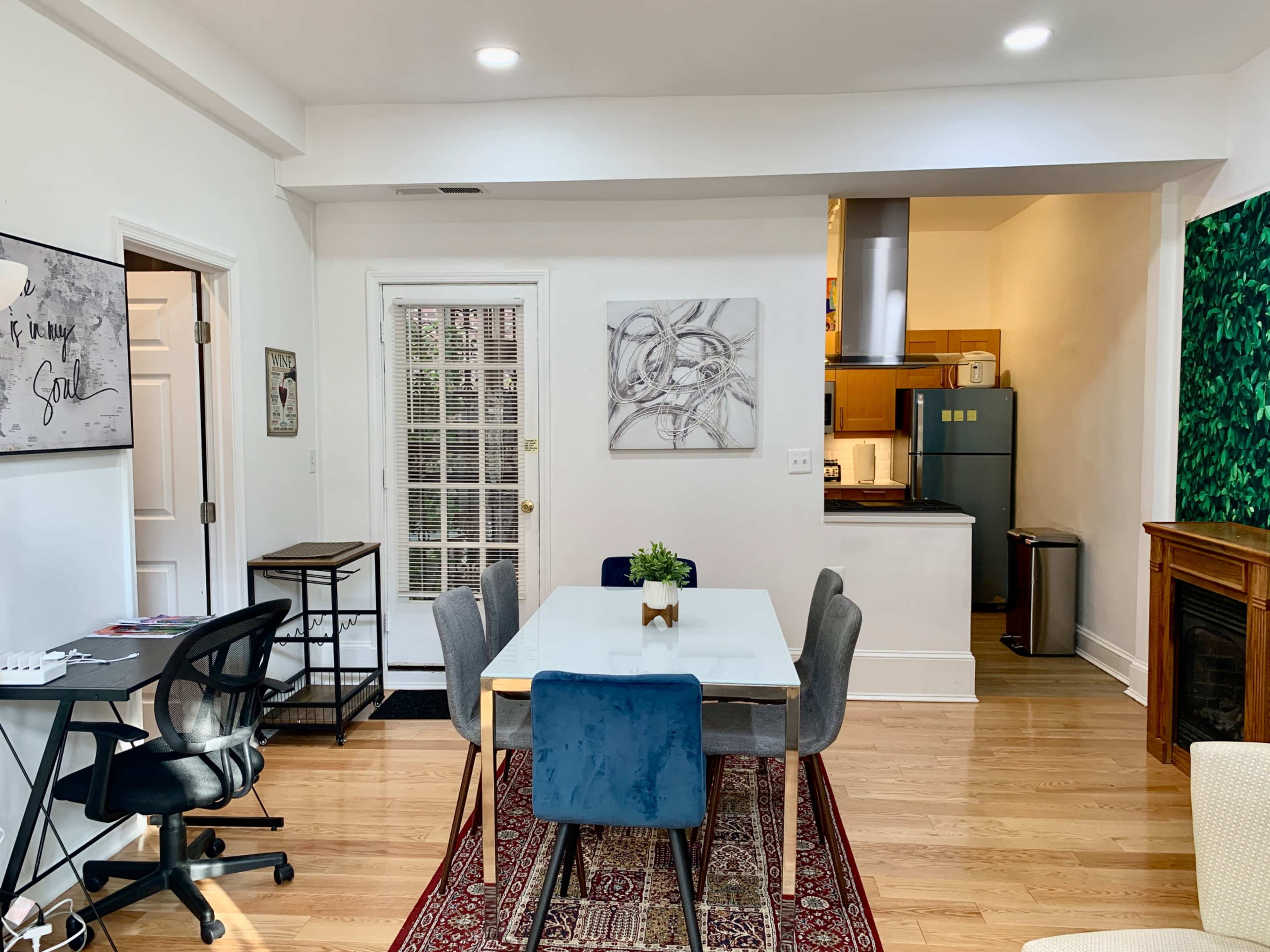 A dining area with a rectangular table surrounded by chairs, leading into a kitchen with appliances visible in the background.