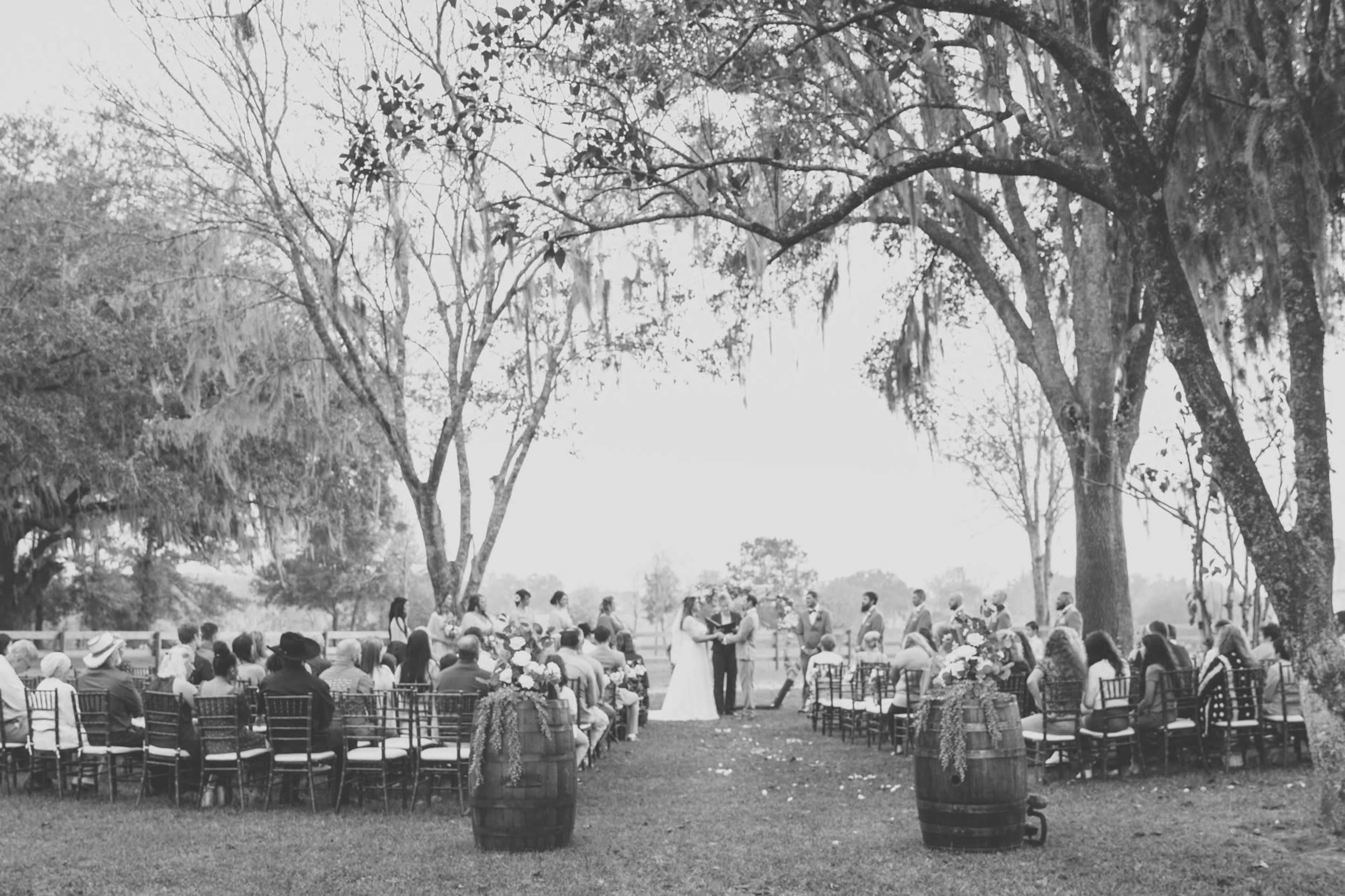 A wedding ceremony is set up outdoors with rows of chairs arranged under trees and a couple standing at the altar.