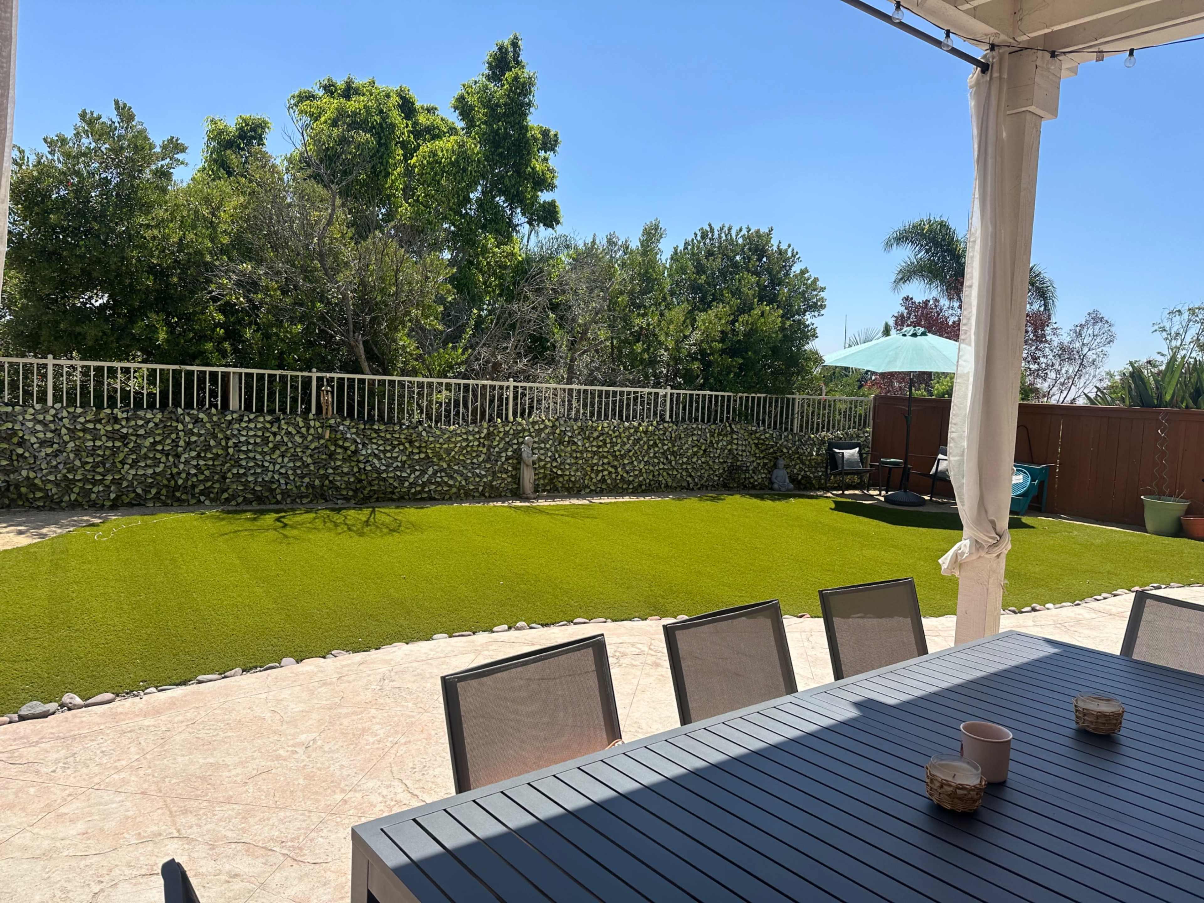 A patio table is set up under a covered area, overlooking a well-maintained green lawn and surrounding trees in a residential backyard.