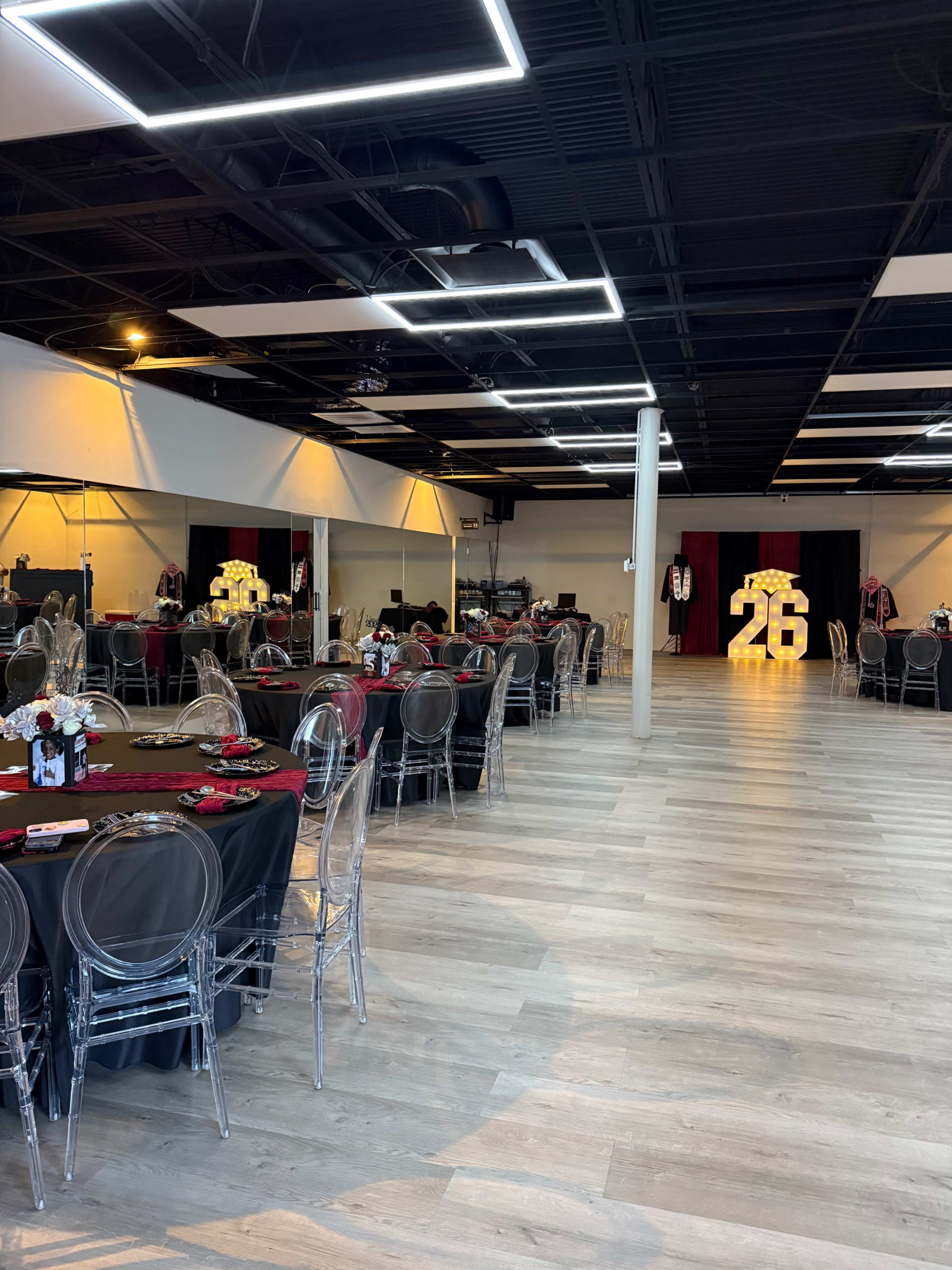 The image shows a spacious event hall set up with round tables covered in black and red tablecloths, featuring clear ghost chairs, and illuminated numbers "26" against a decorated backdrop.