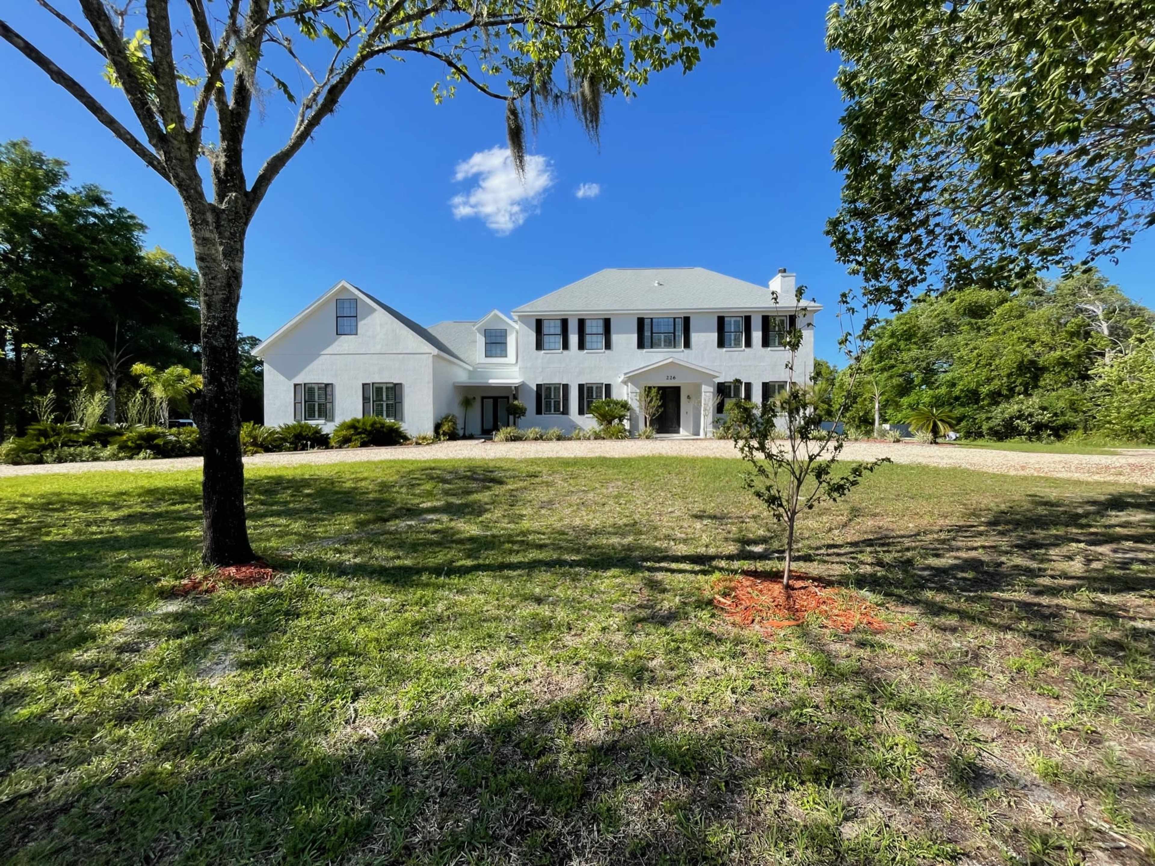 A two-story white house with black shutters is set in a grassy yard surrounded by trees.