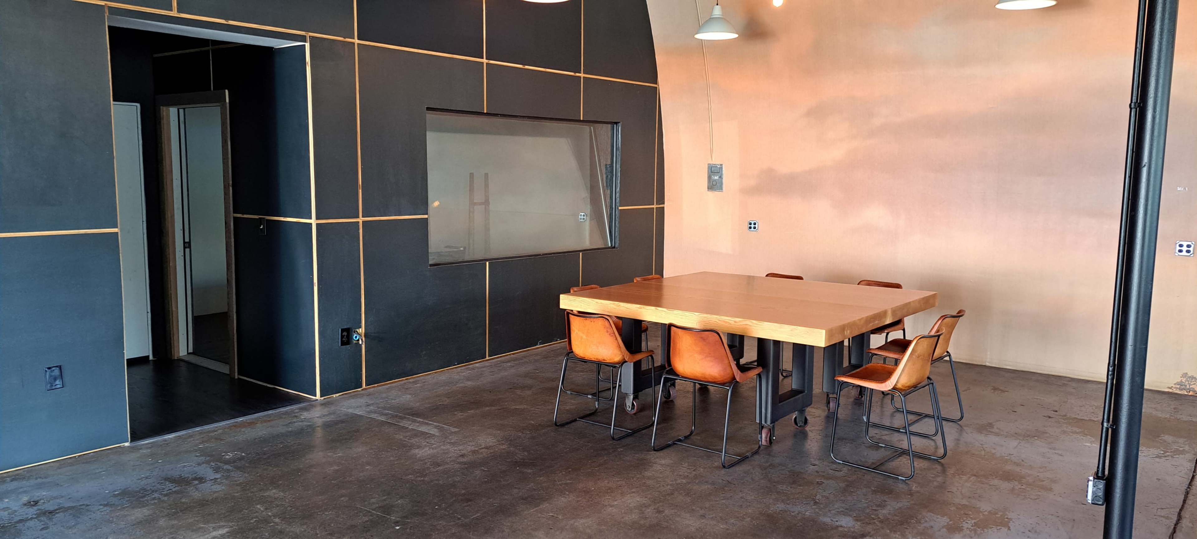 The image shows a modern dining area featuring a large wooden table surrounded by six chairs, with a partially visible adjacent room and dark walls.