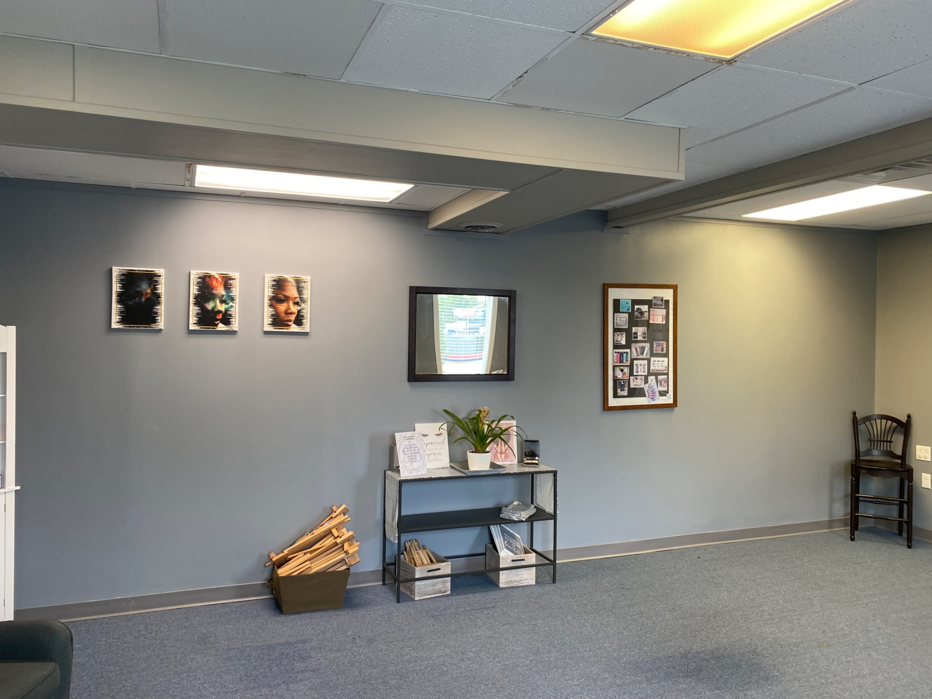 A room with a gray wall featuring four framed photographs, a mirror, a display board with various pictures, and a shelf holding items, alongside a chair in the corner.