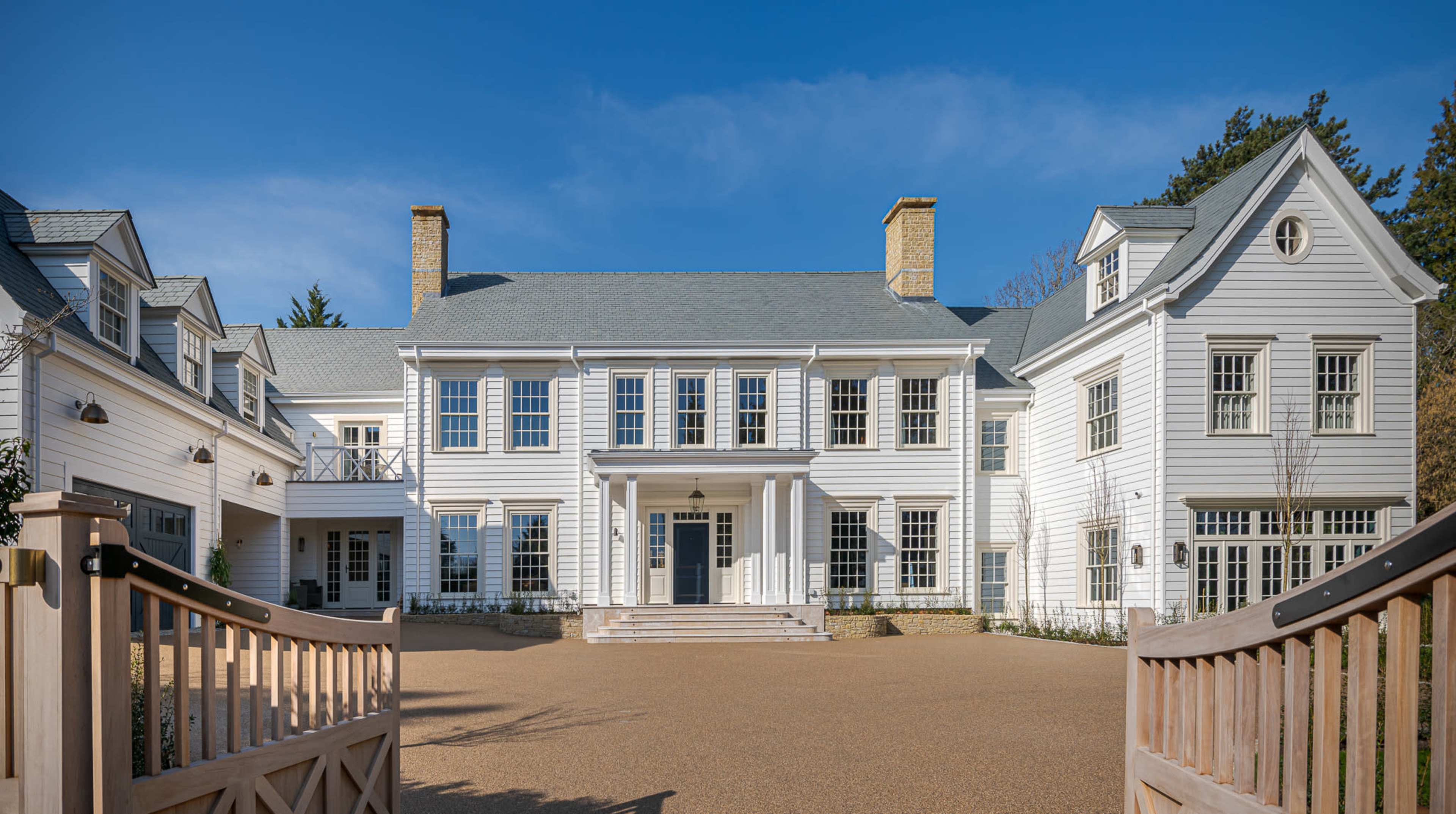 The image depicts a large, white, colonial-style mansion with multiple gables and a broad entrance, surrounded by a gravel driveway.