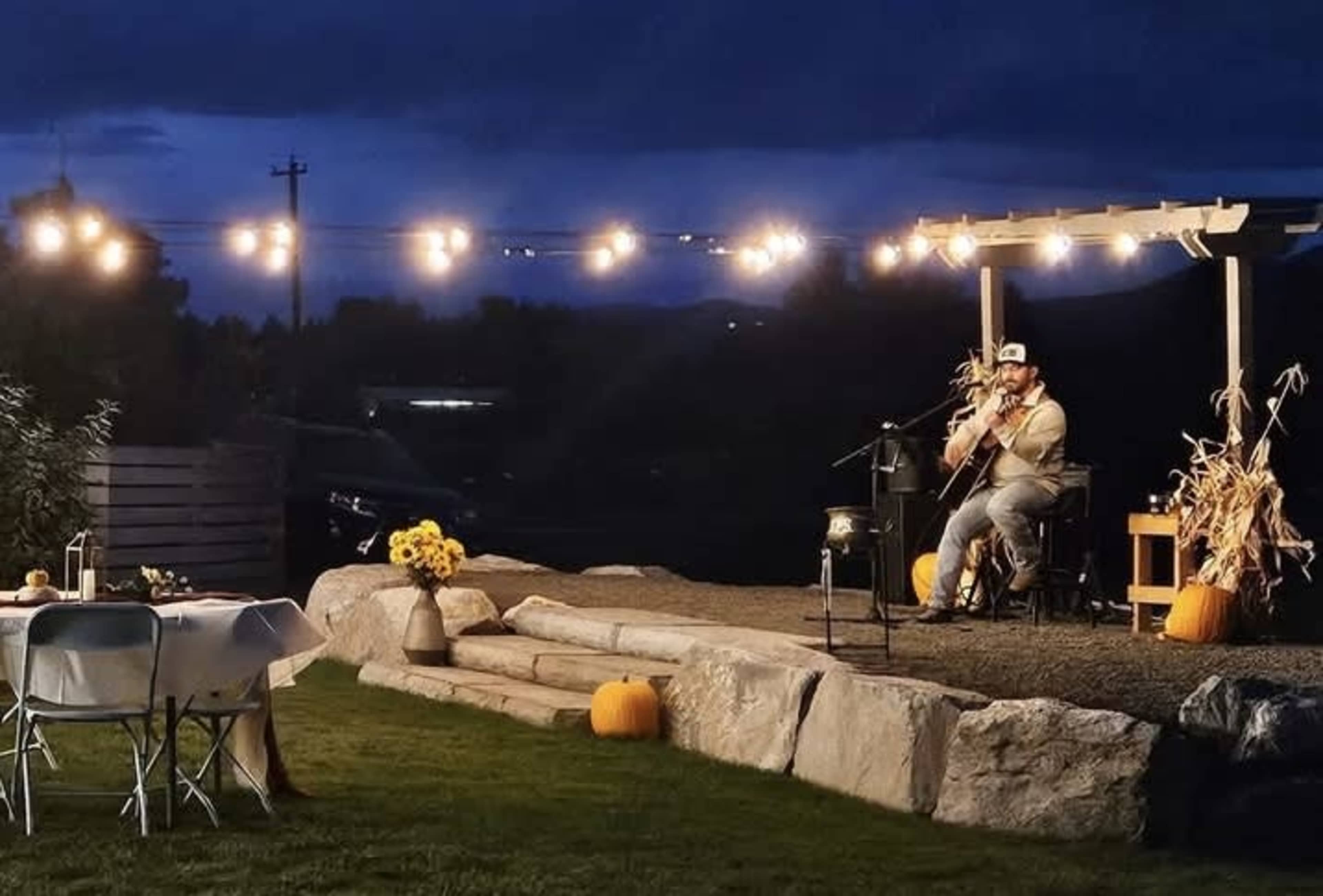 A musician plays guitar on a stage illuminated by string lights, surrounded by pumpkins and a set dining area in the evening.