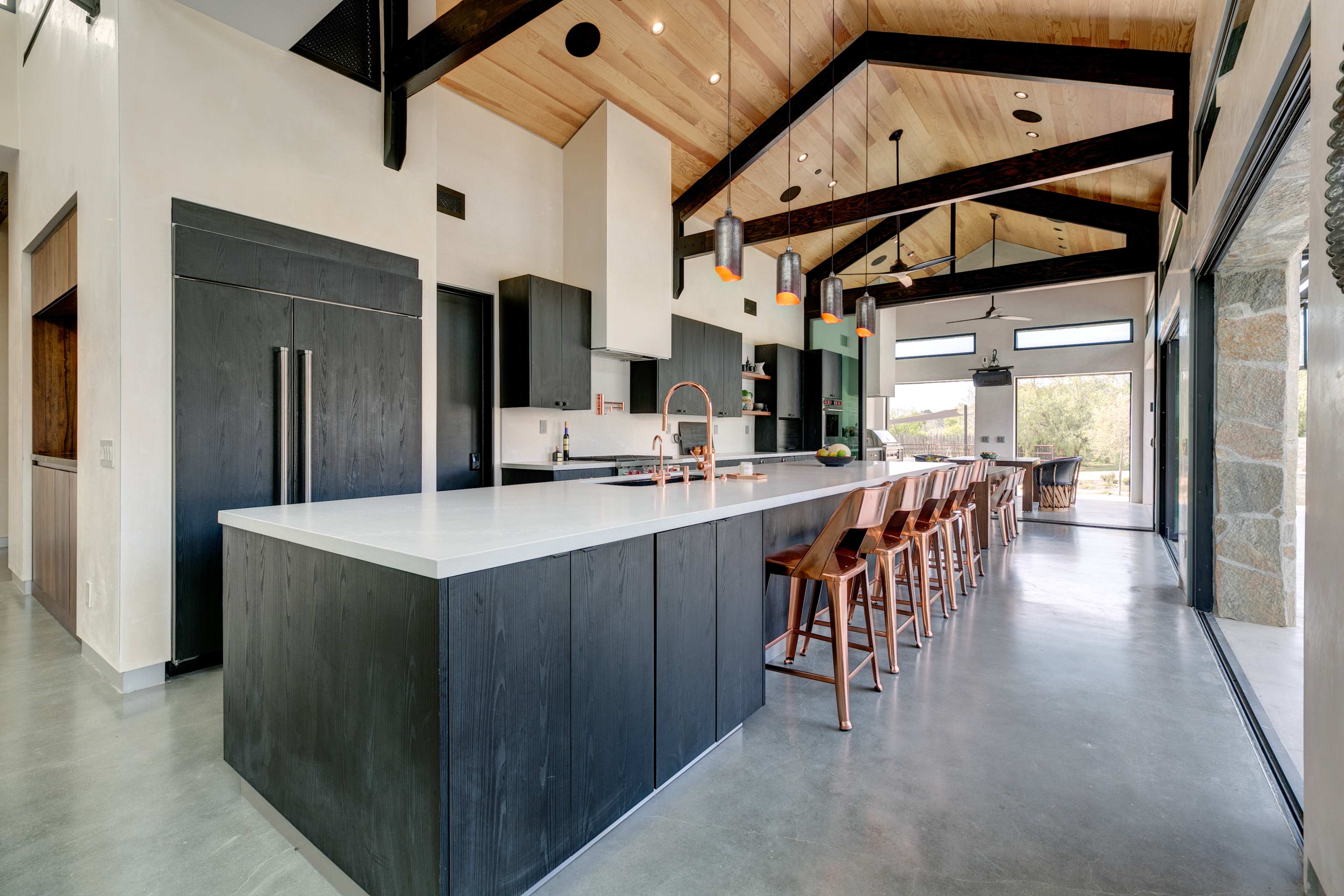A modern kitchen features a large central island with a white countertop, black cabinetry, and wooden bar stools under a vaulted ceiling with exposed beams.