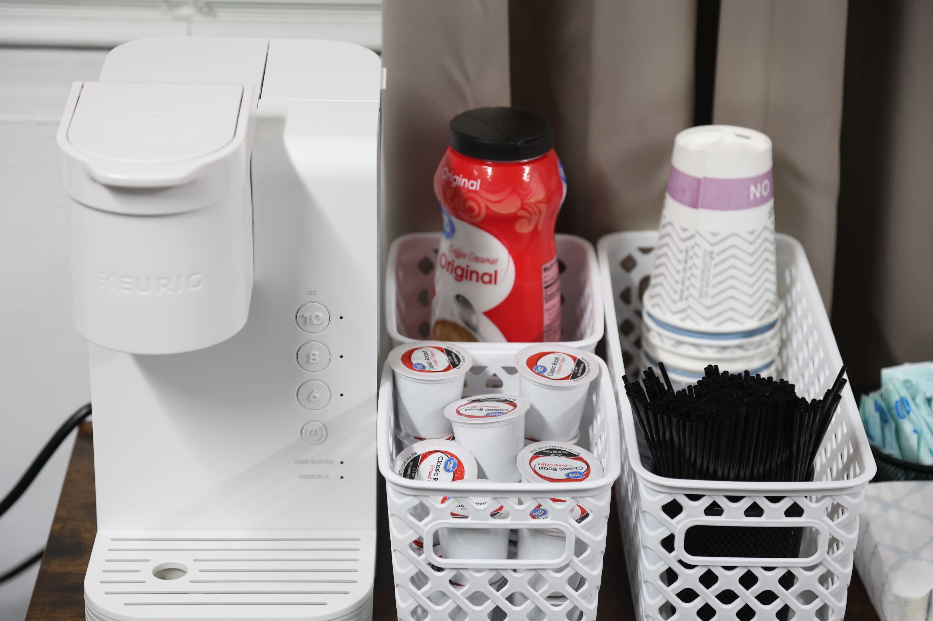 The image shows a Keurig coffee maker alongside containers of coffee pods, a bottle of creamer, paper cups, and plastic stirrers organized on a countertop.