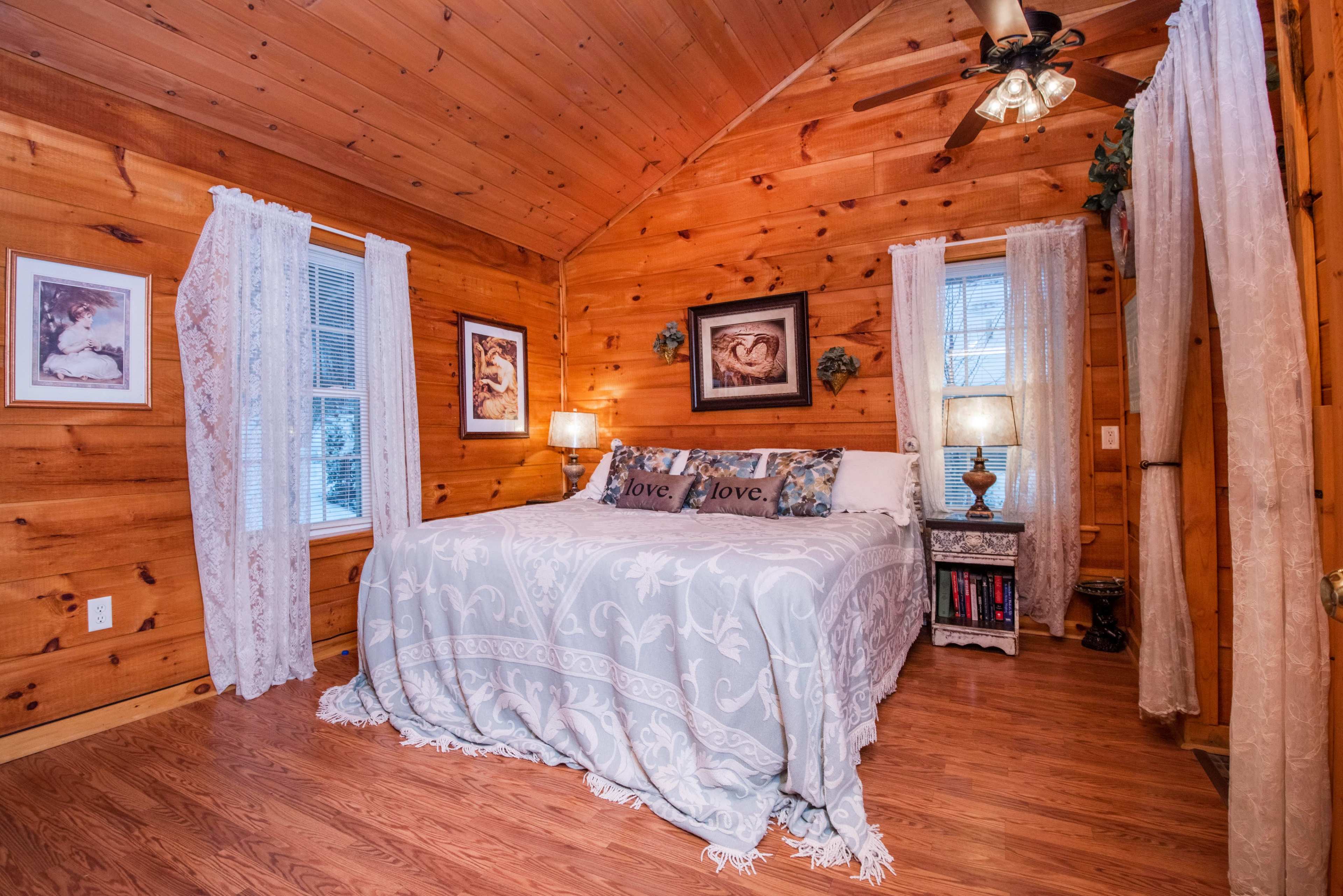 The room features a wooden interior with a bed dressed in a light gray coverlet, two windows dressed with white curtains, and framed artwork on the walls.
