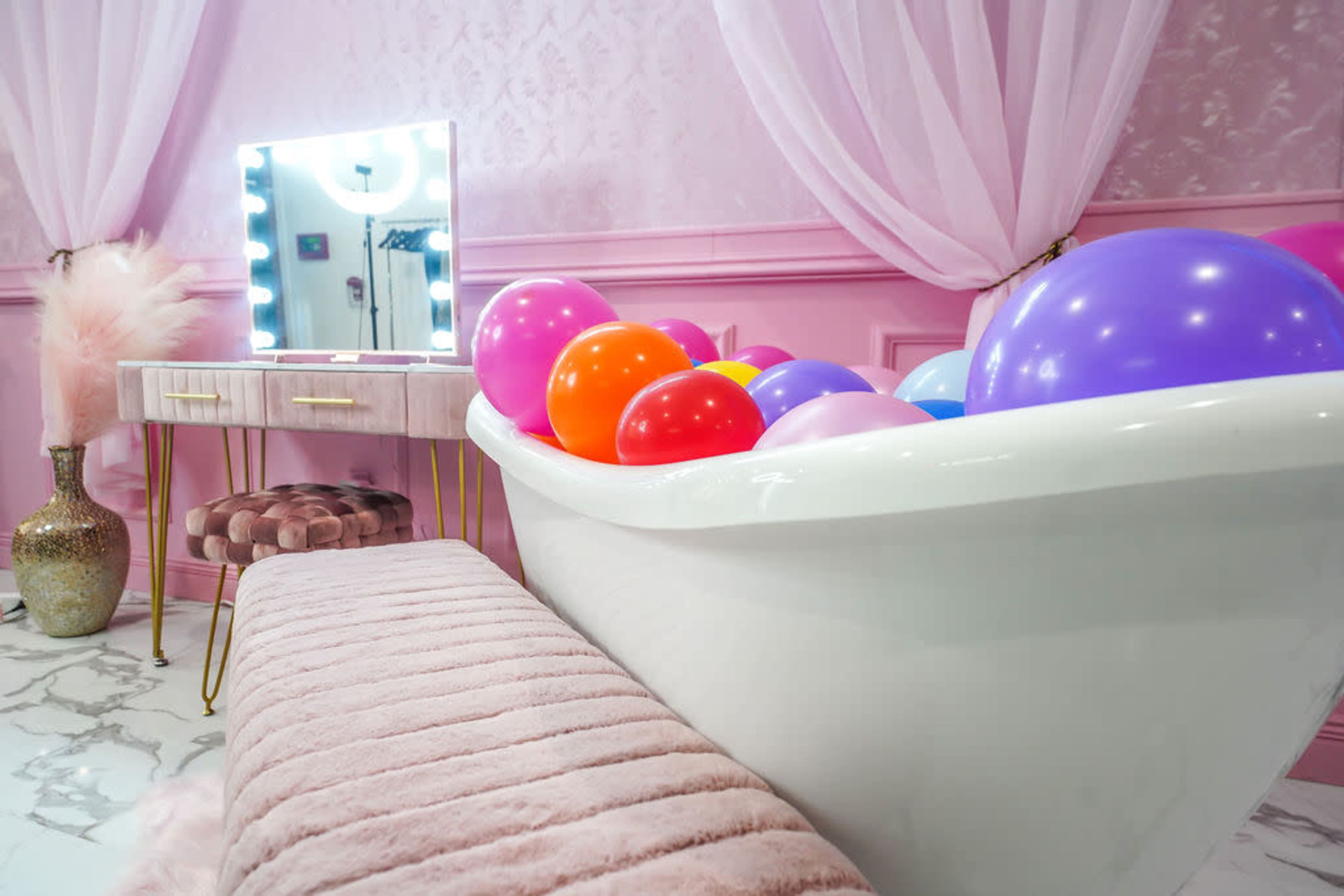 A white bathtub filled with colorful balloons is positioned next to a vanity with a mirror and a pink, plush bench in a elegantly decorated room.