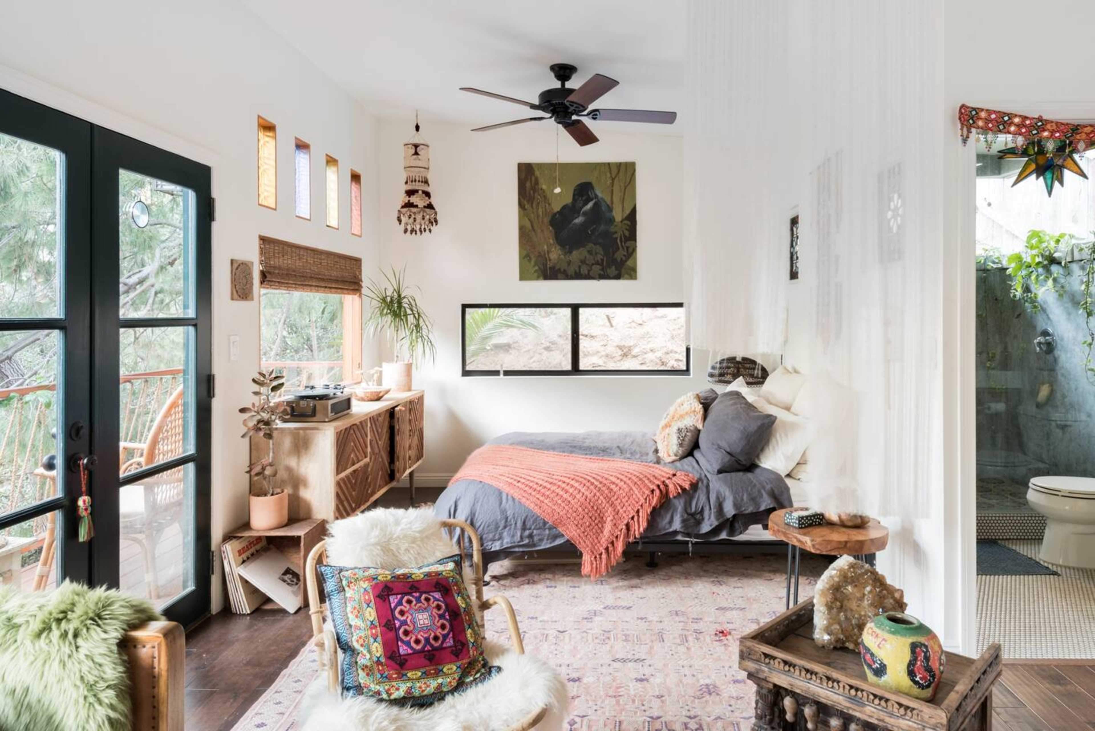 A cozy bedroom features a large bed, natural light from windows, and earthy decor elements including plants and textiles.