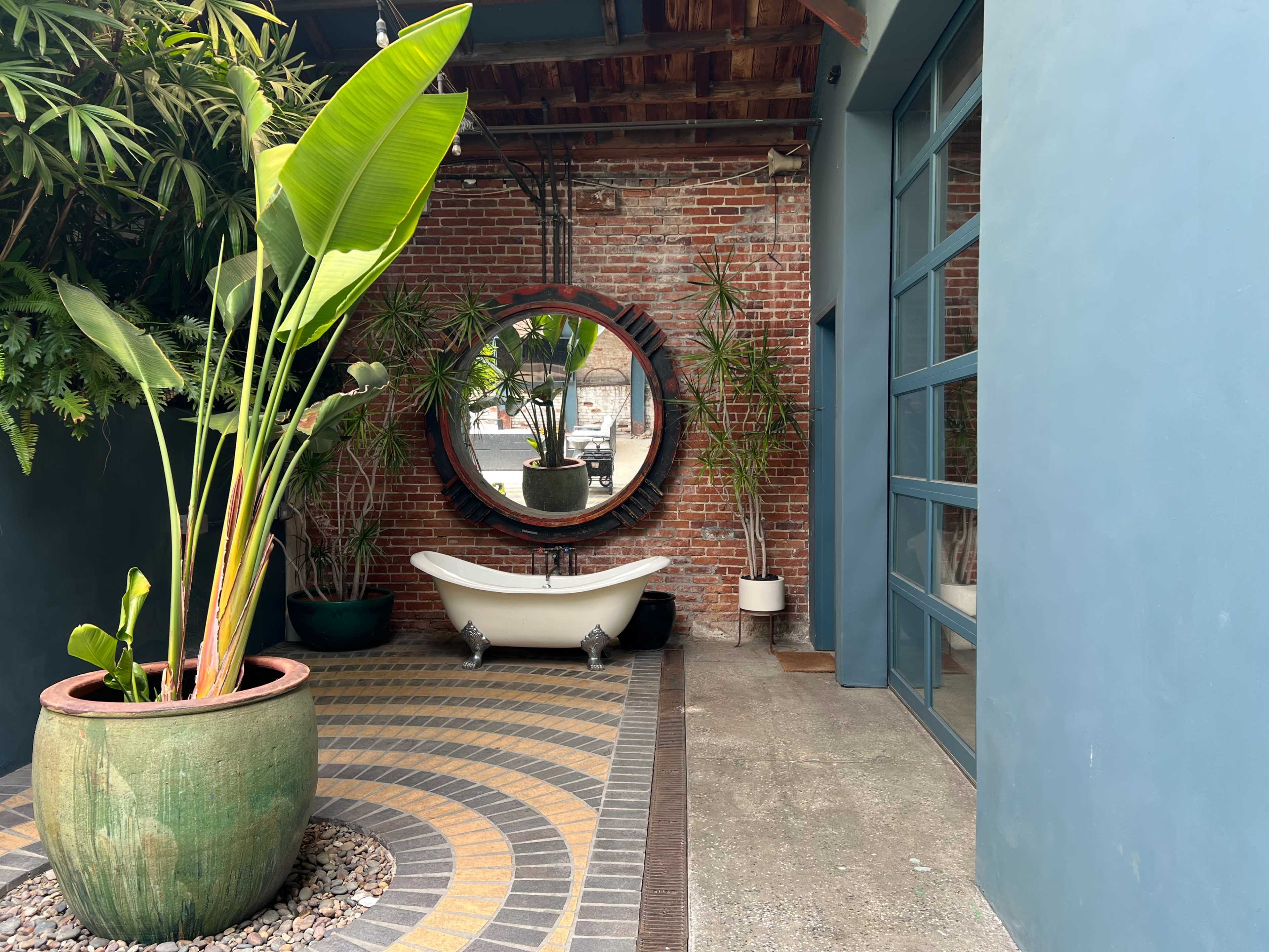 The image shows a modern indoor space with a round mirror, potted plants, a vintage bathtub, and a brick wall.