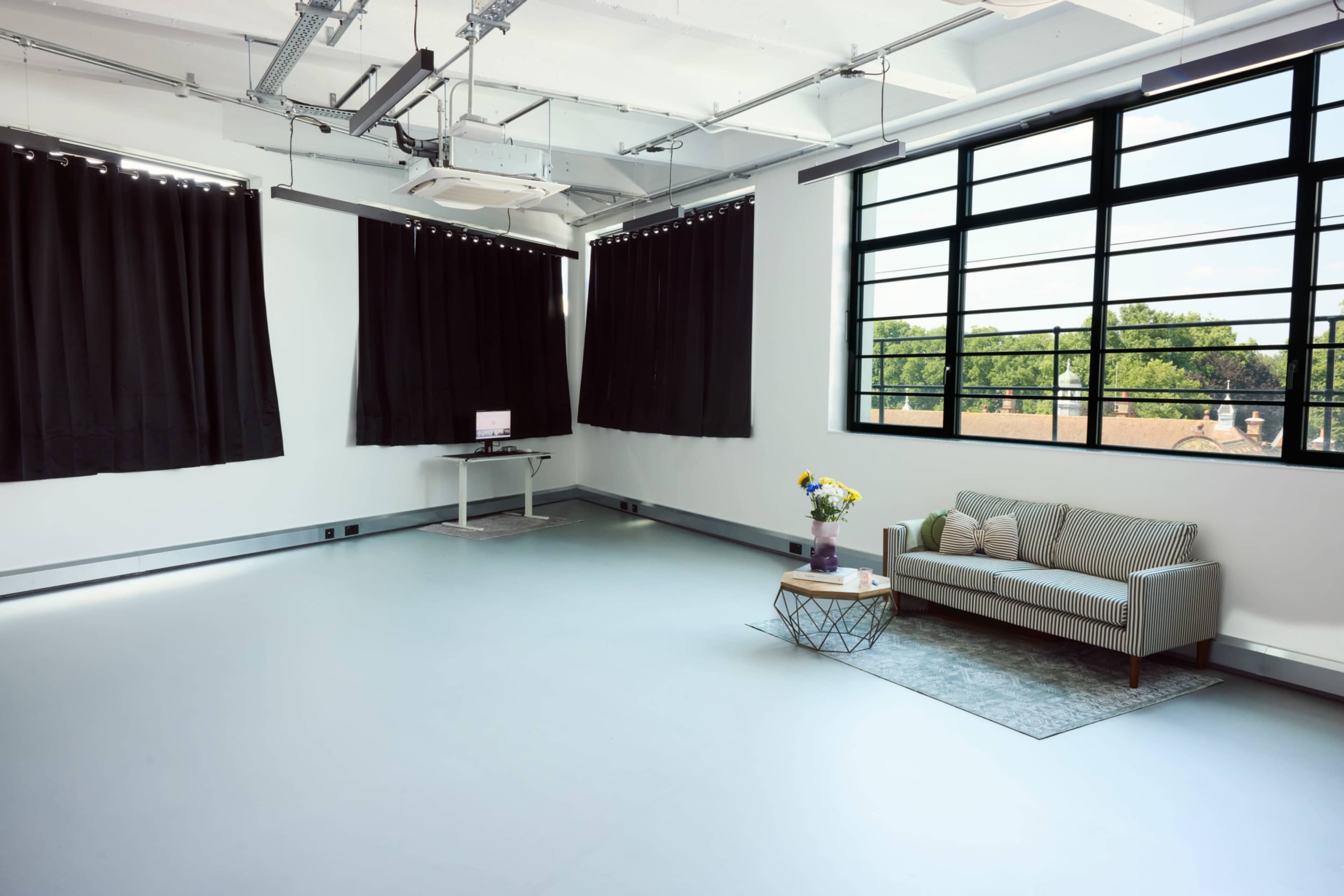 The image shows a spacious, minimalist room with large windows, a gray sofa, a coffee table, and dark curtains.