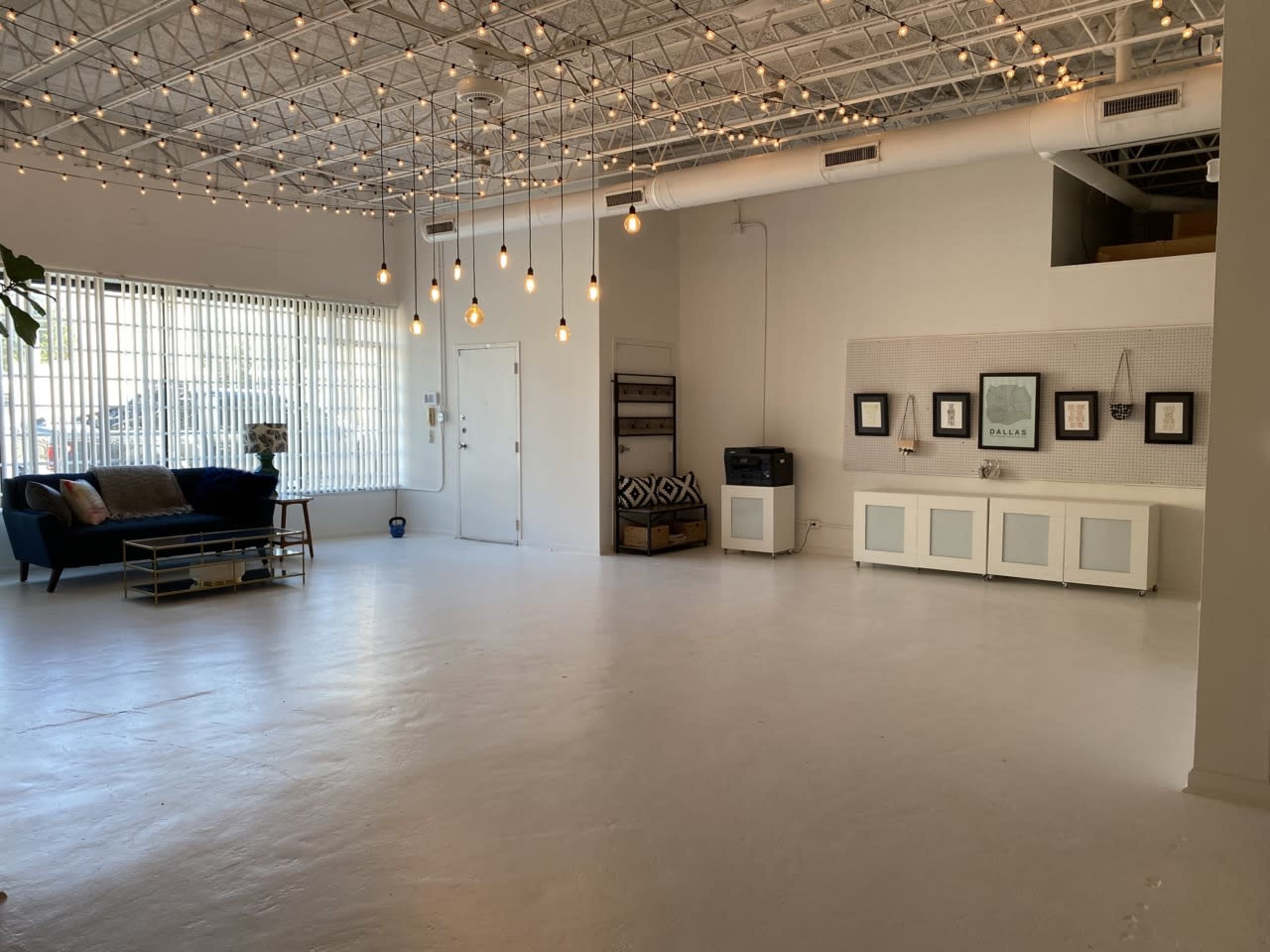 The image shows a spacious, well-lit interior with white walls, a concrete floor, a seating area with a blue couch, and decorative pendant lights hanging from the ceiling.