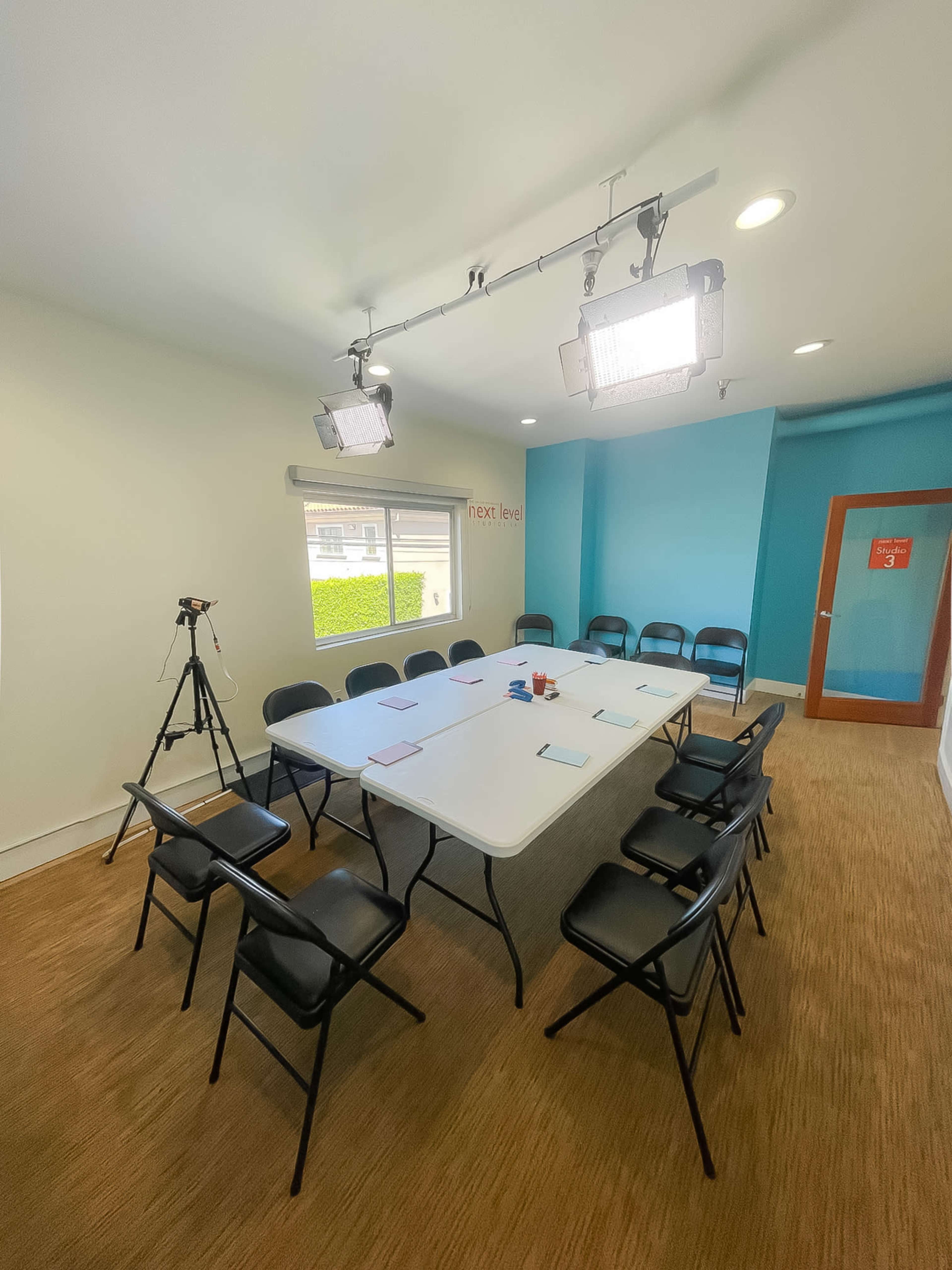 A meeting room is set up with a large table surrounded by black chairs, equipped with a camera on a tripod and bright overhead lights.