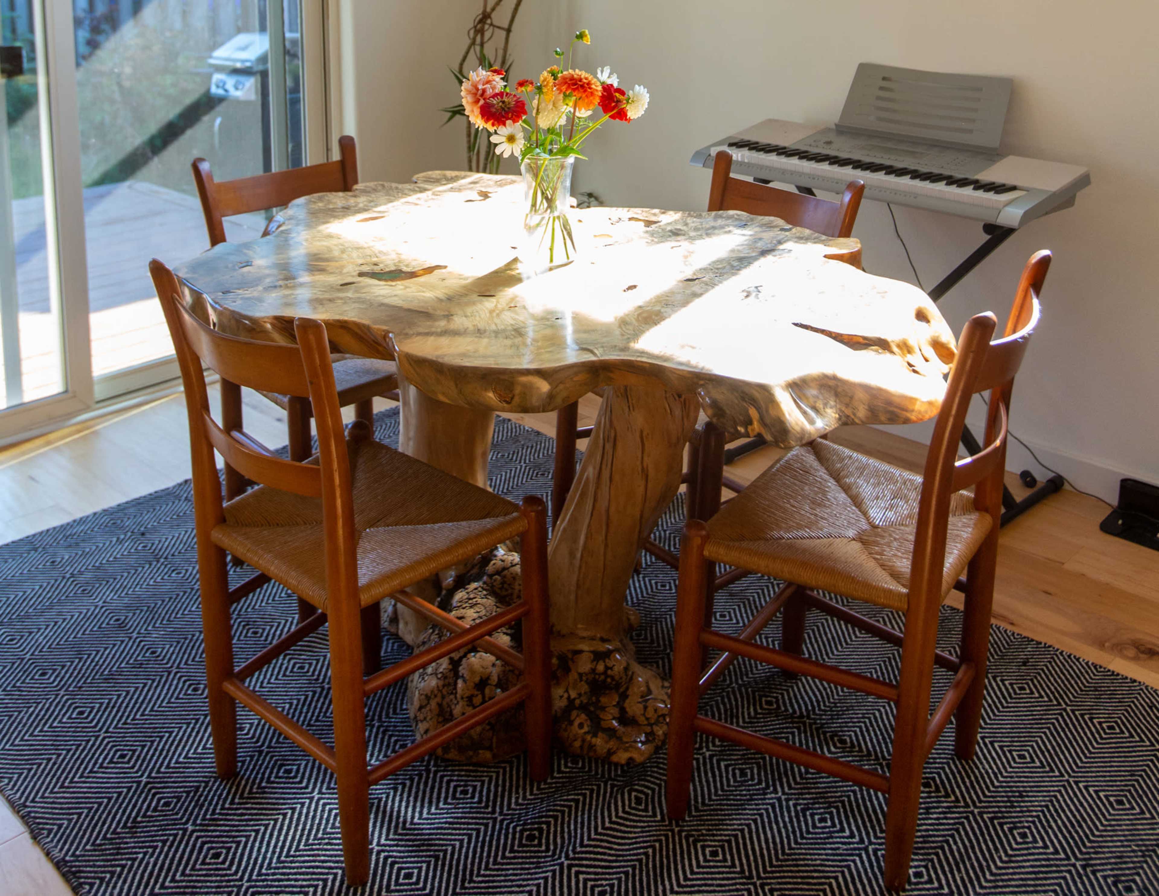 A wooden dining table with a natural edge is surrounded by four wooden chairs, and a vase of flowers sits atop the table in a room with a keyboard in the background.
