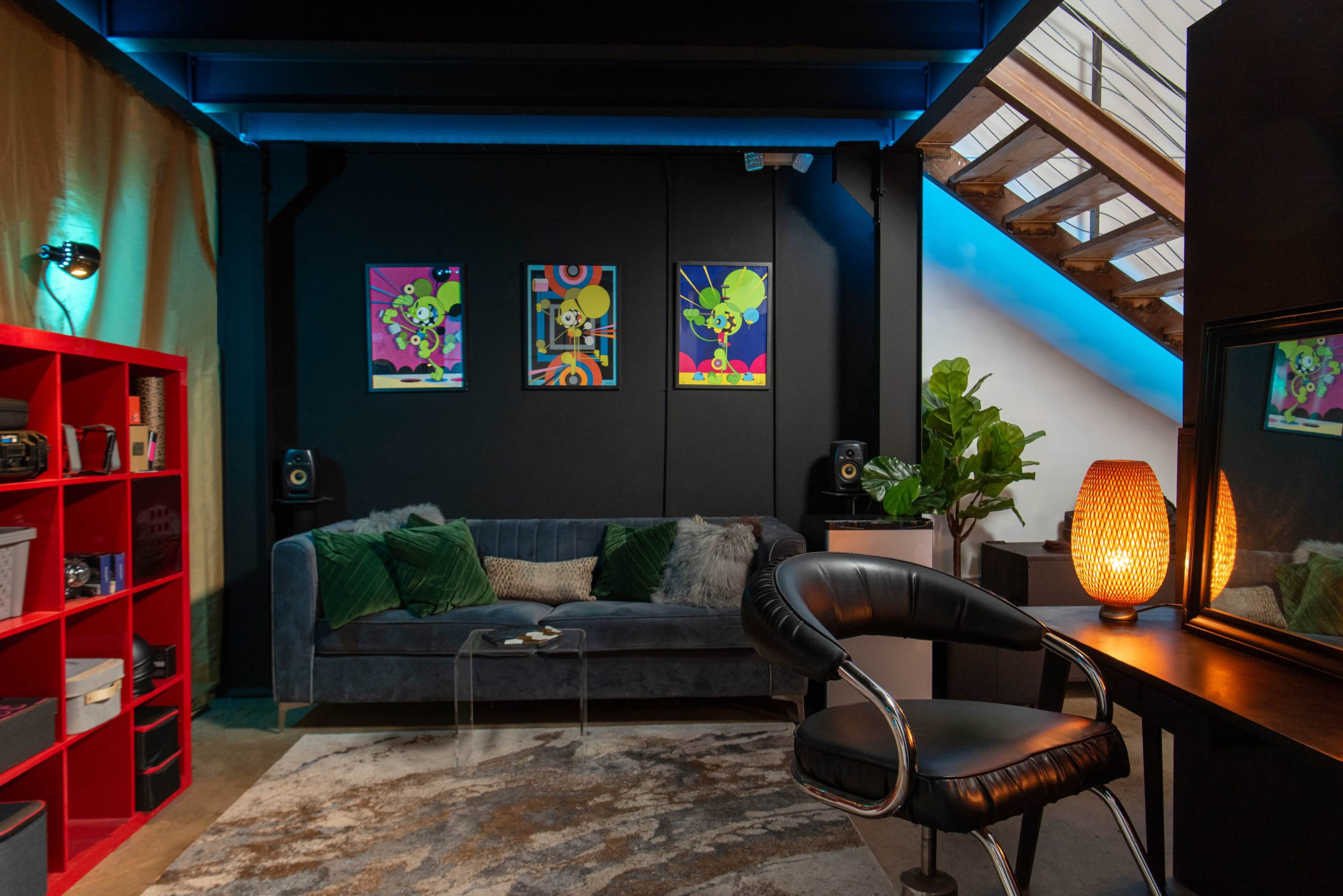 The image shows a modern living space featuring a velvet couch, a bookshelf, and colorful artwork on the walls, with a staircase in the background.