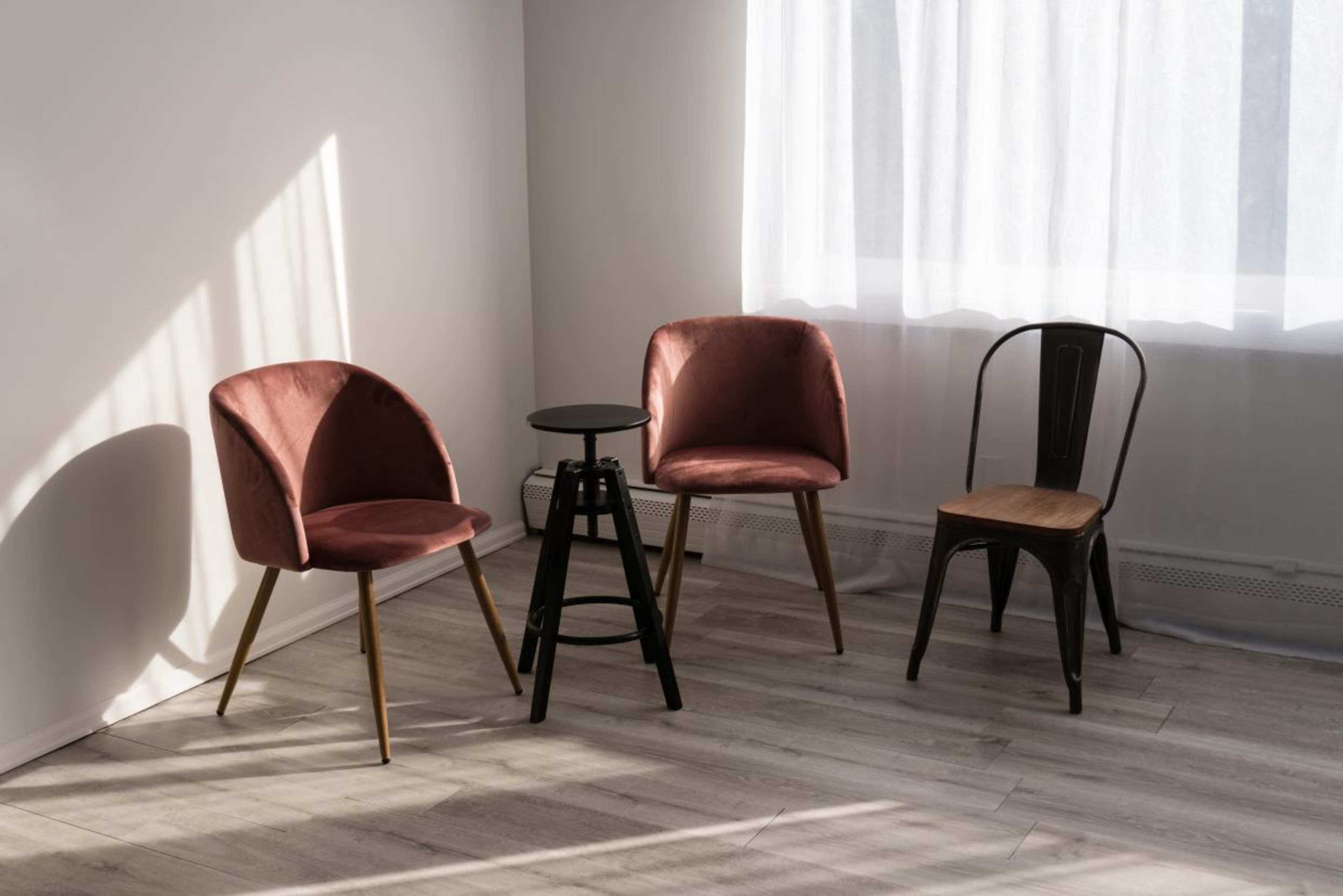 Two pink upholstered chairs and a black stool positioned near a window with sheer curtains, accompanied by a black metal chair.