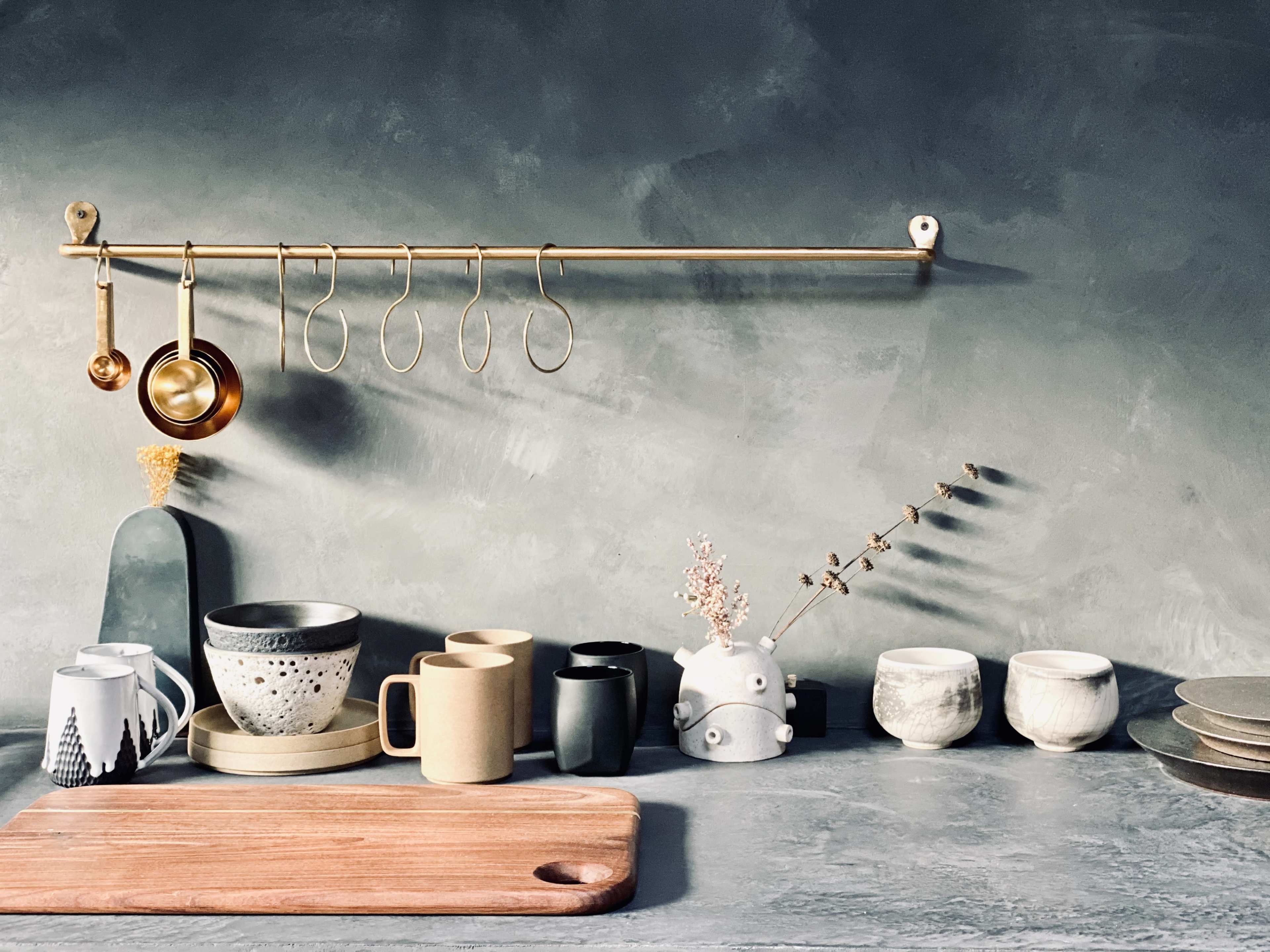 A collection of various ceramic mugs and decorative items is displayed on a grey countertop beneath a brass rack with hanging utensils.