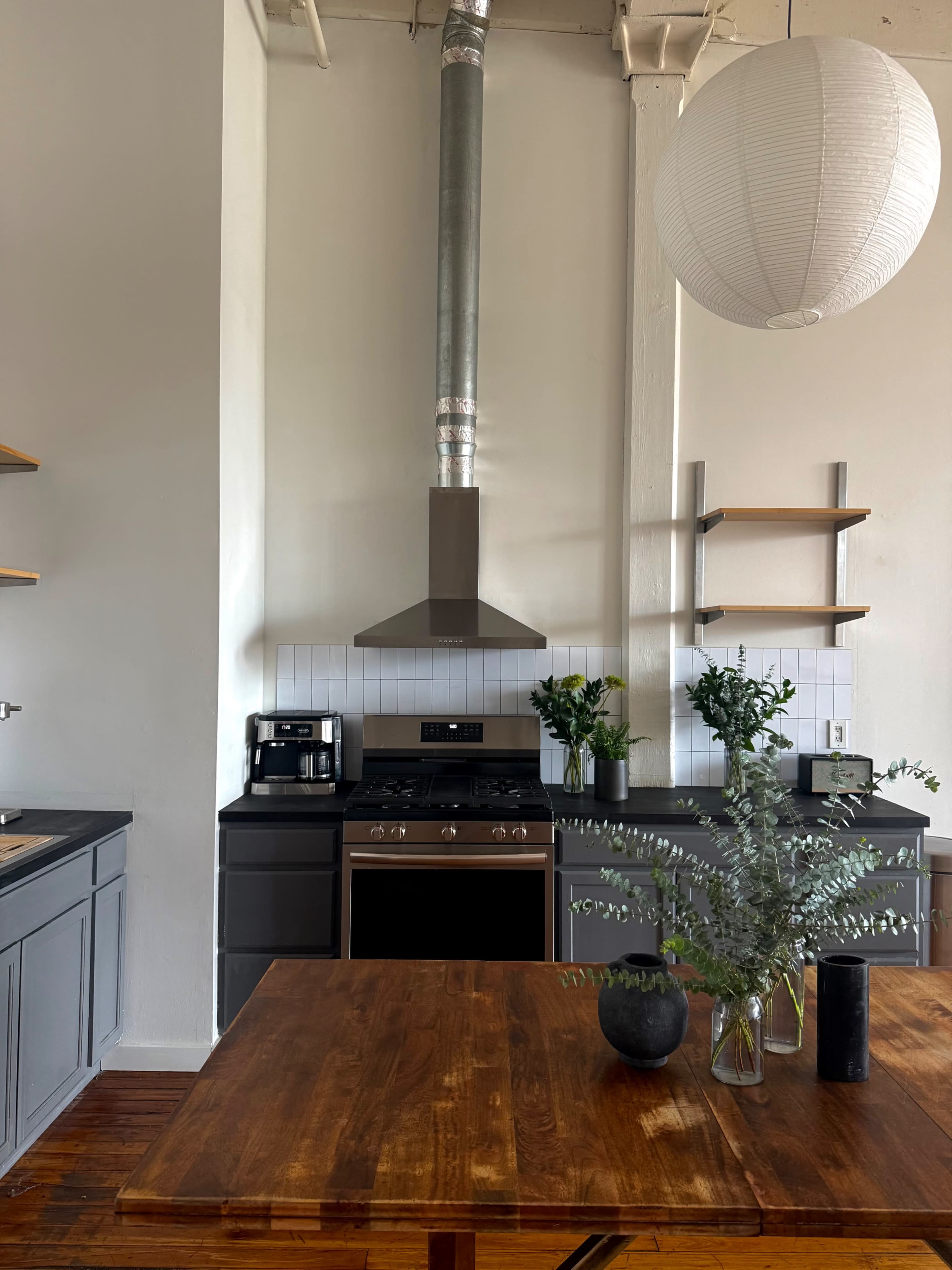 The image shows a modern kitchen featuring a stainless steel range hood, open shelving, and a wooden dining table with a vase of greenery.