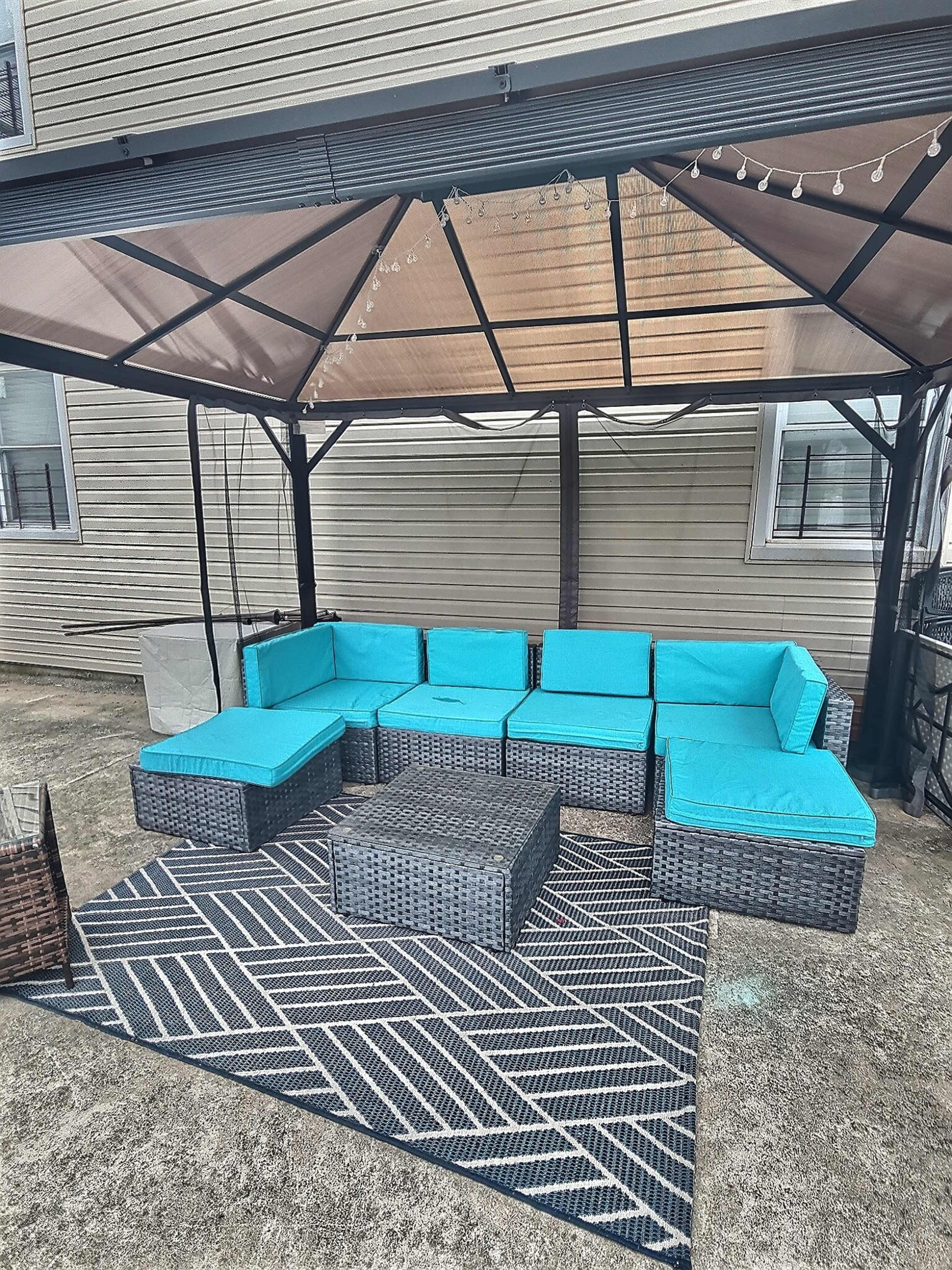 A patio area features a turquoise cushioned sectional sofa under a canopy, accompanied by a geometric patterned rug and a square coffee table.