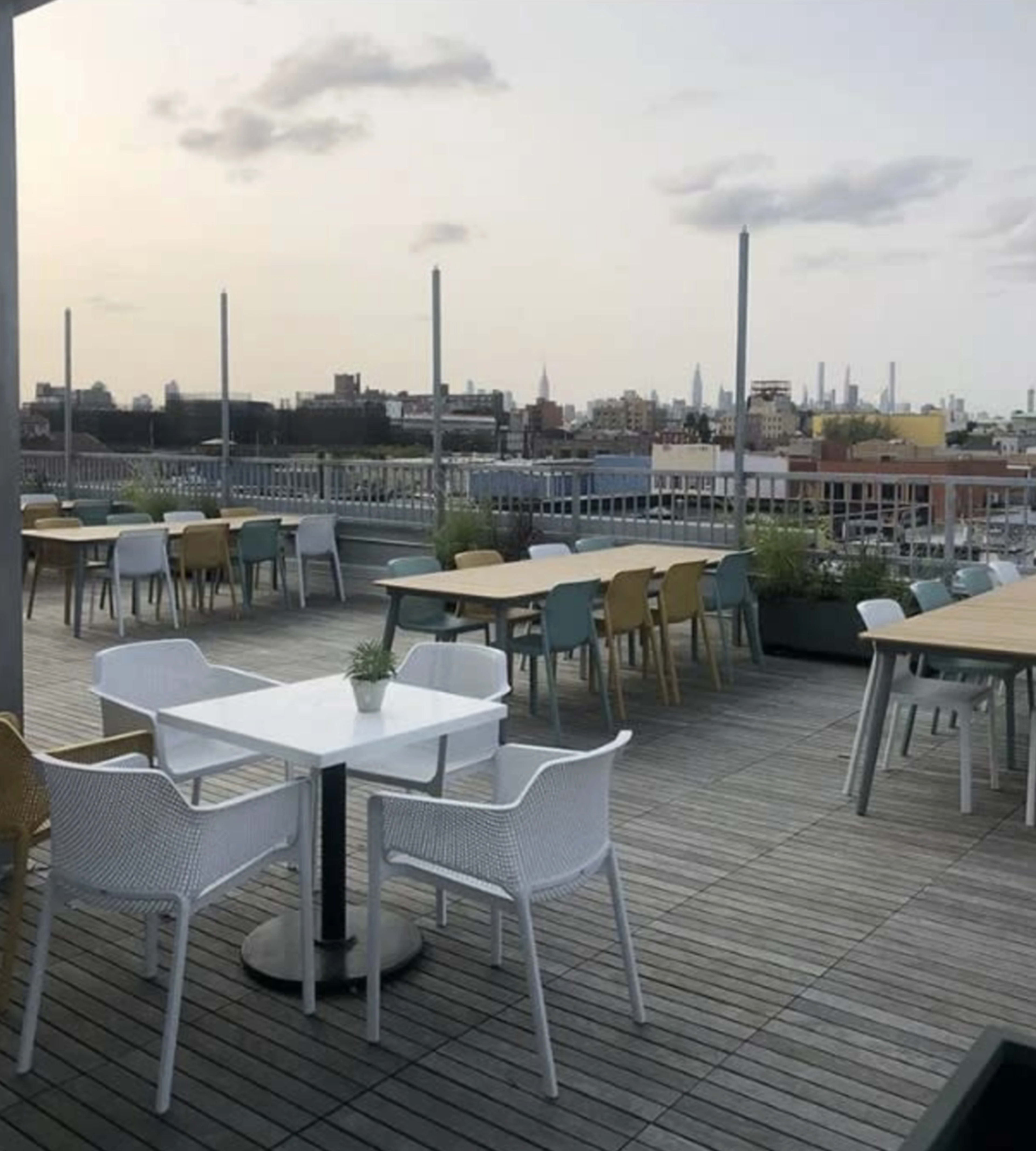 The image shows a rooftop terrace with various tables and chairs arranged on a wooden deck, overlooking a city skyline in the background.