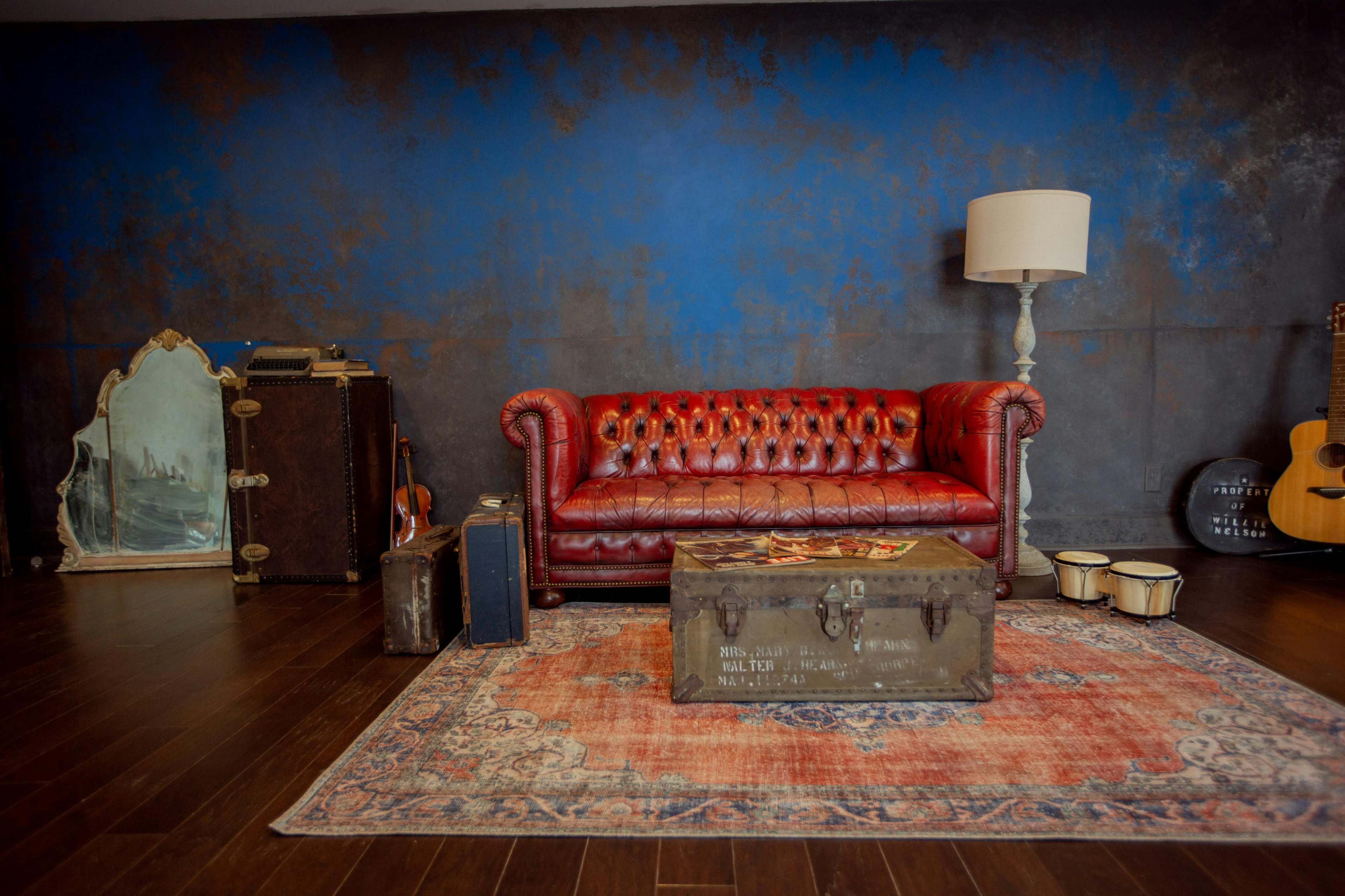 The image depicts a vintage-style room with a red leather chesterfield sofa, a wooden coffee table, an ornate mirror, stacked luggage, a lamp, a guitar, and a set of drums on a patterned rug.