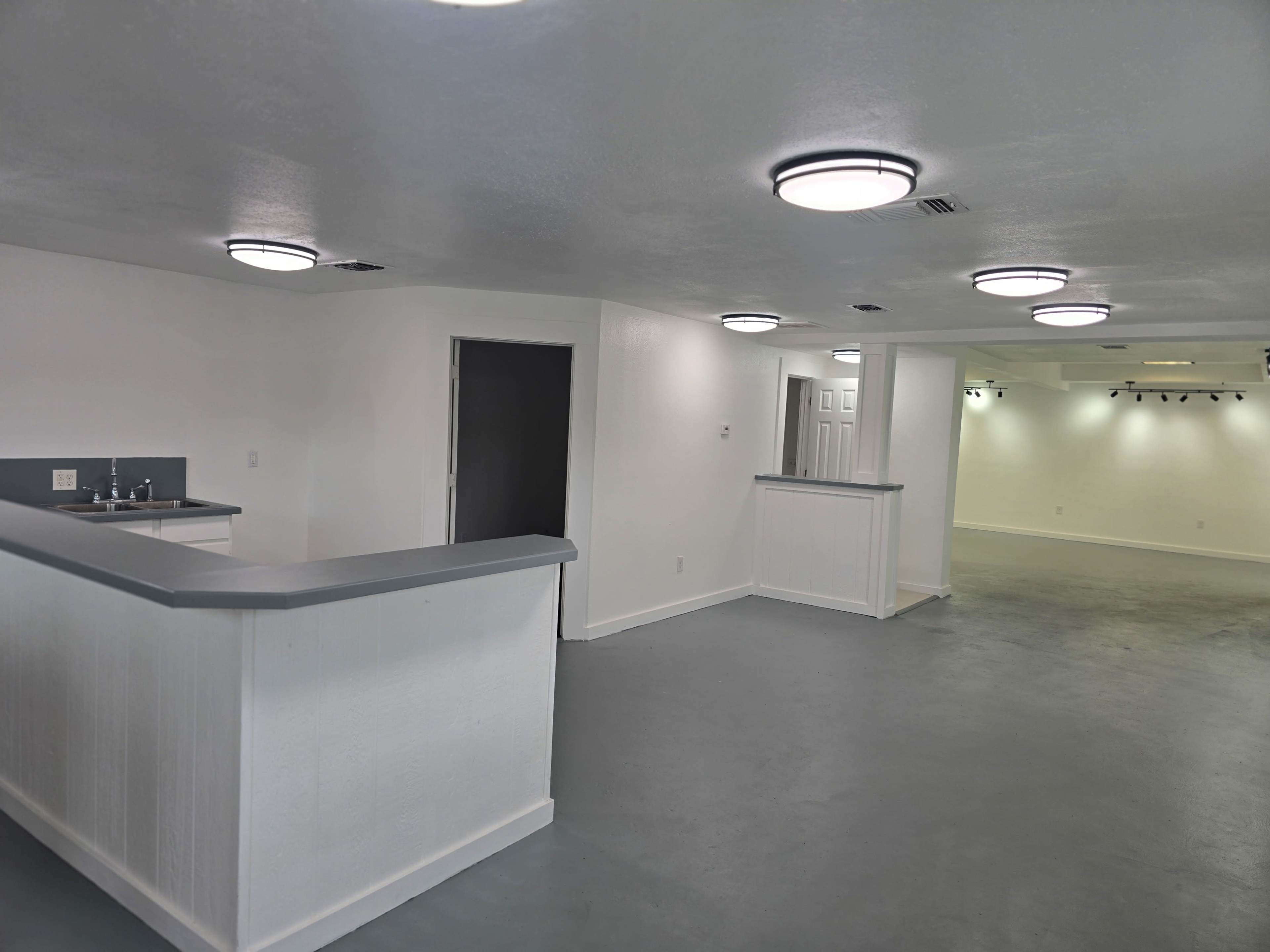 The image shows a spacious, well-lit interior with a gray floor, white walls, and modern ceiling lights, featuring a small kitchenette area on the left and an empty area for various uses ahead.