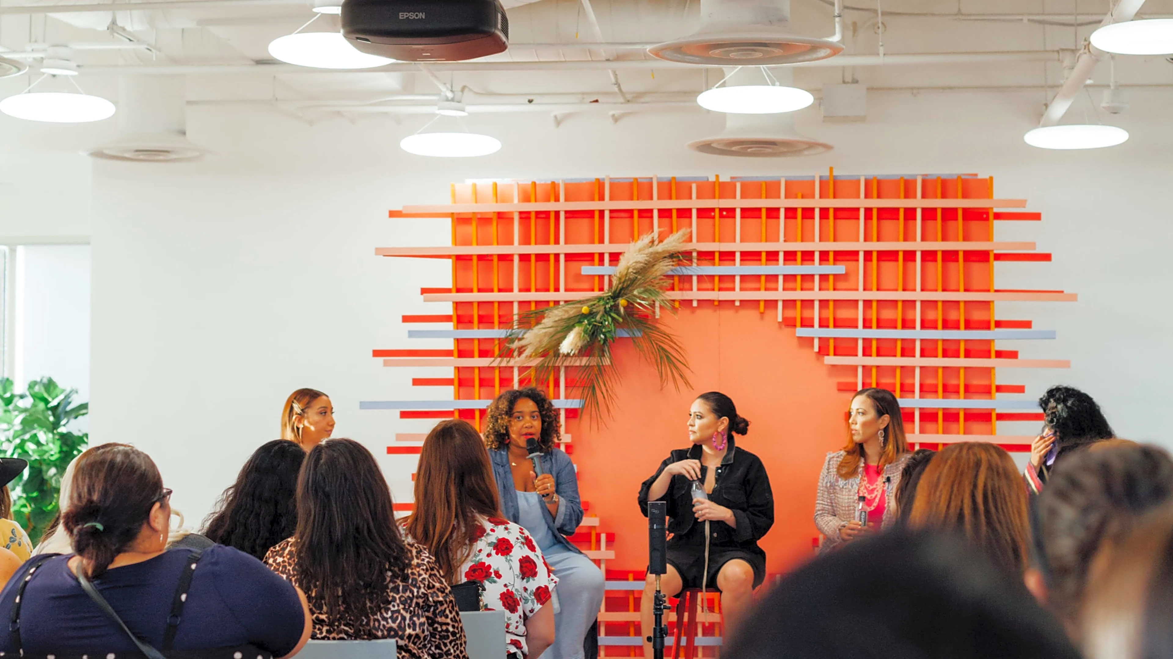 A panel discussion takes place in a brightly lit room with attendees seated in front of four speakers against a colorful backdrop.