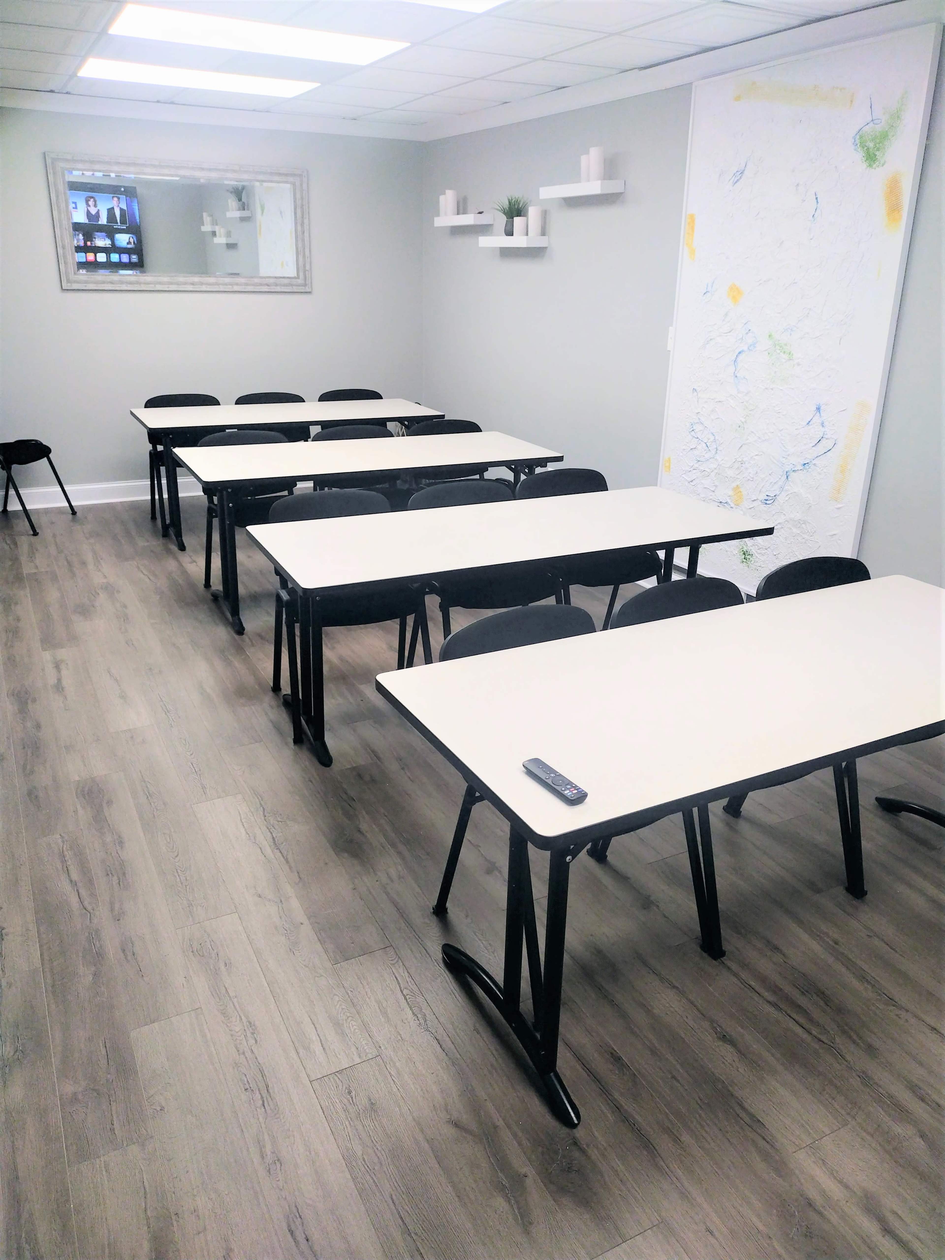 A classroom setup features multiple rows of tables and chairs arranged neatly, with a wall-mounted screen and a large map on the side.