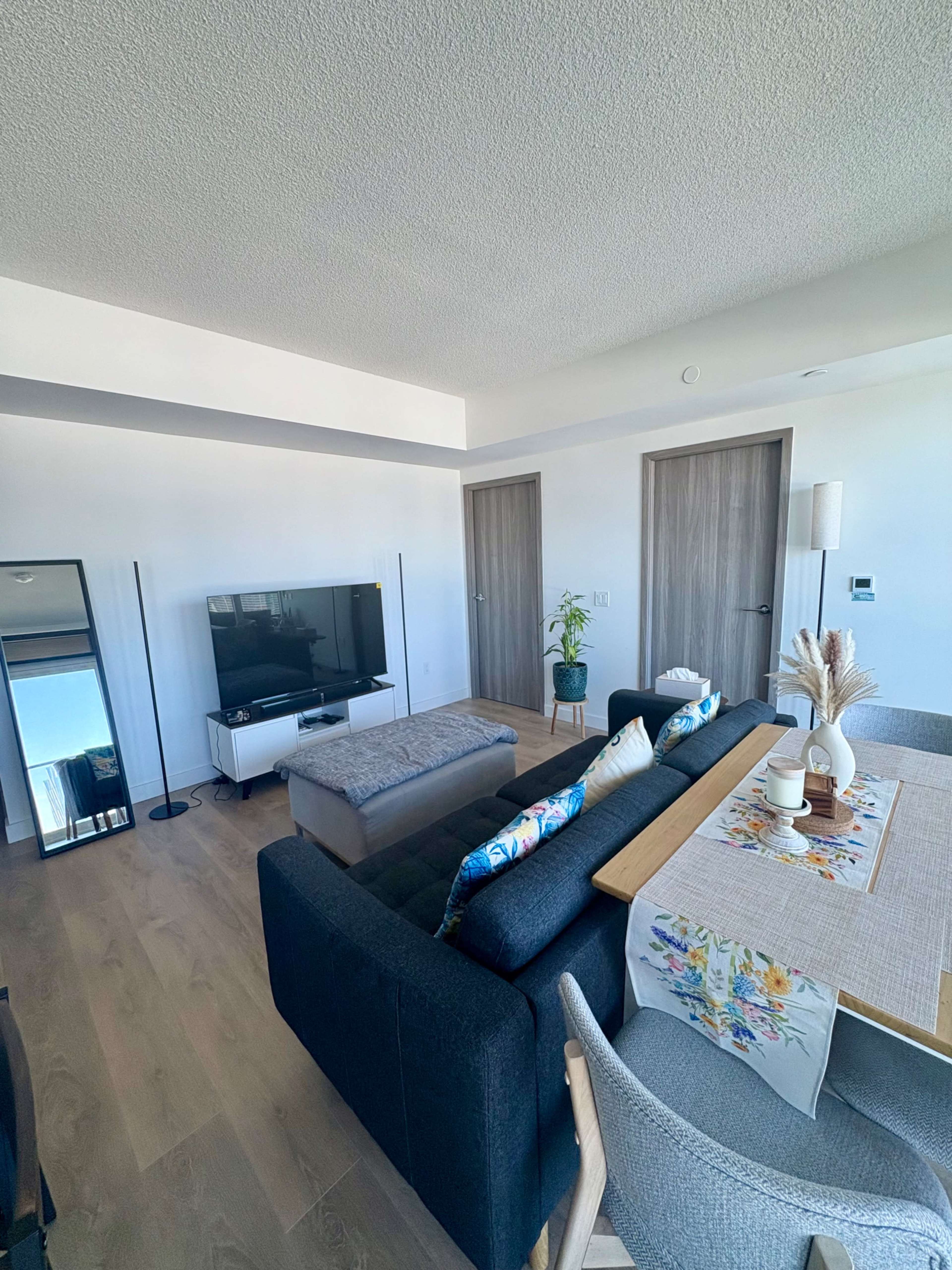 The image shows a modern living room with a dark sofa, a television mounted on the wall, a dining table set for four, and two closed doors leading to other rooms.
