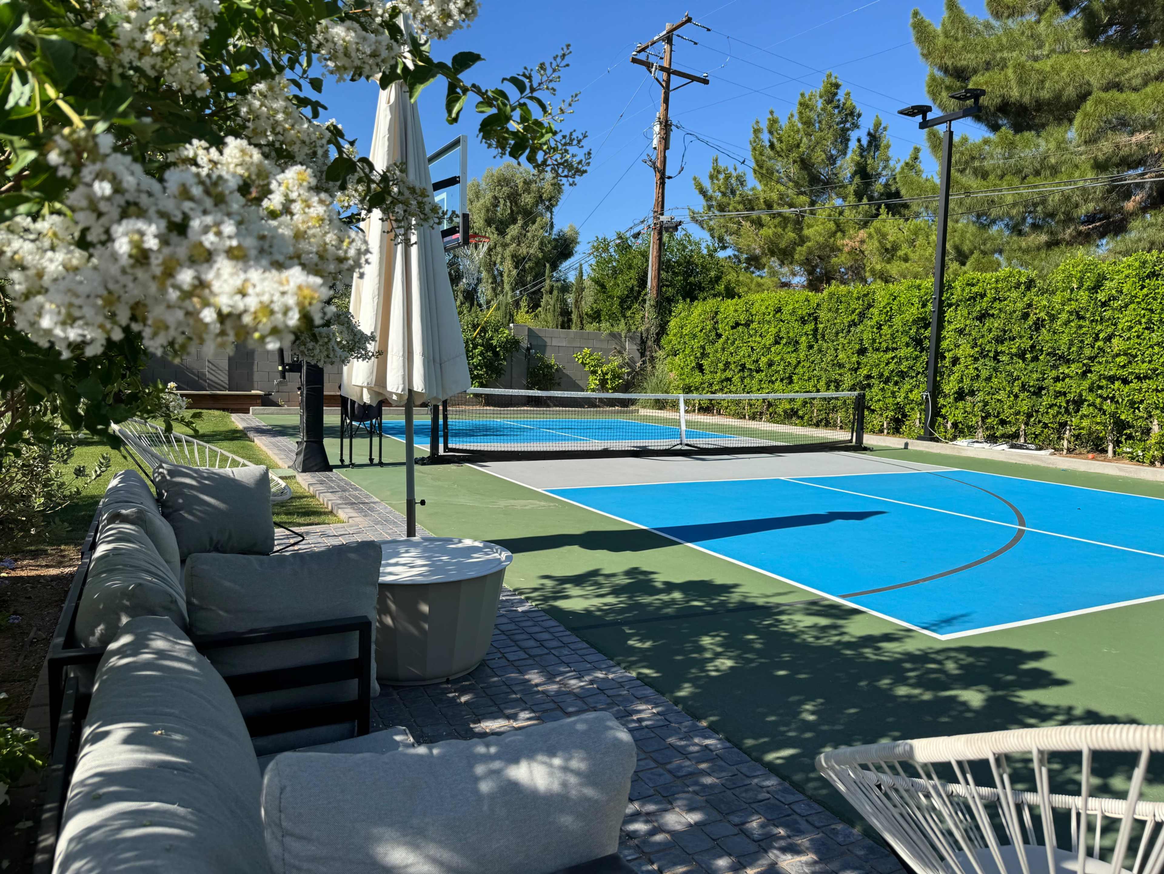 A tennis court surrounded by greenery, with a seating area featuring a couch, table, and umbrellas in the foreground.