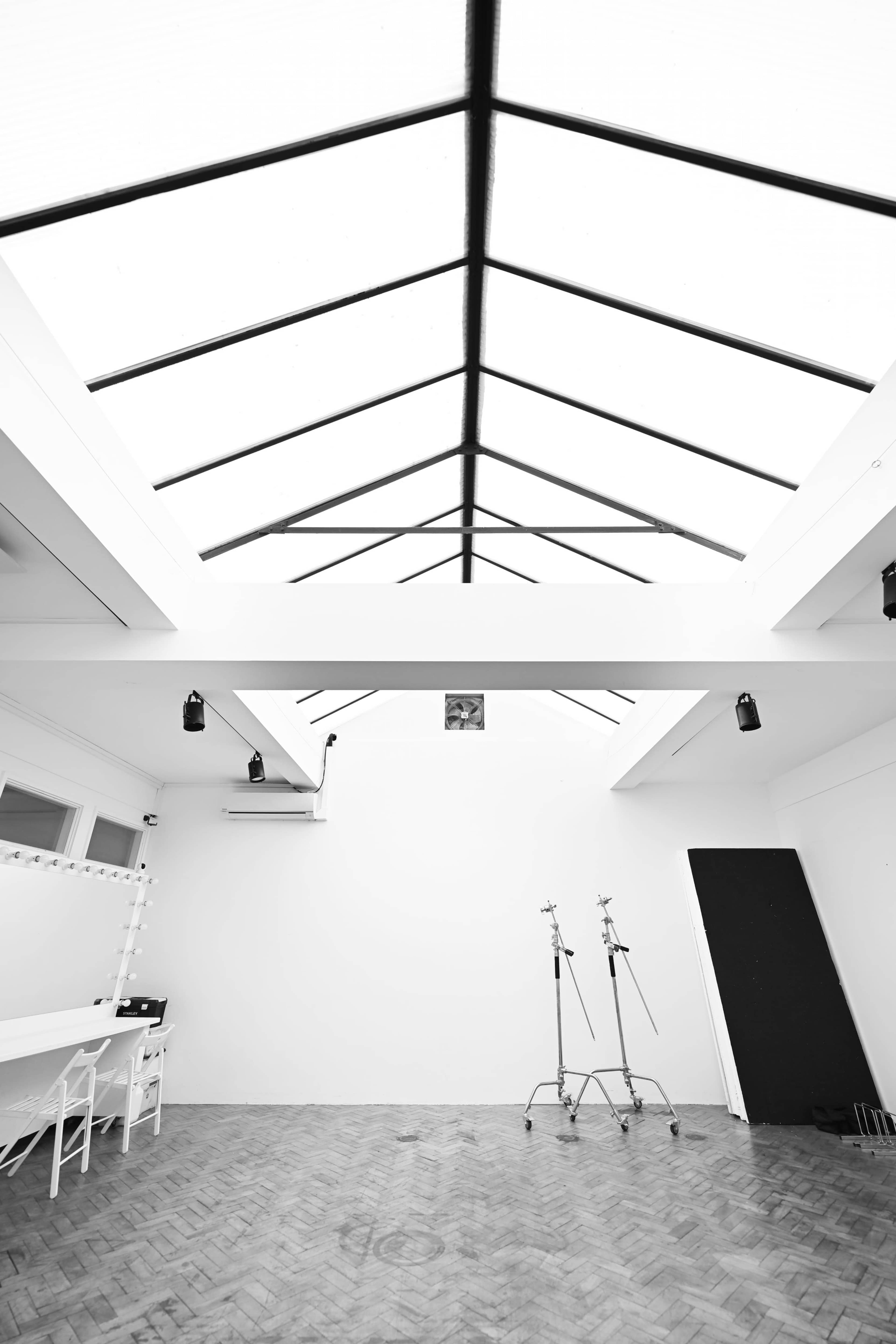 The image shows a minimalistic room with a high, skylighted ceiling, white walls, a wooden floor, and two studio light stands.