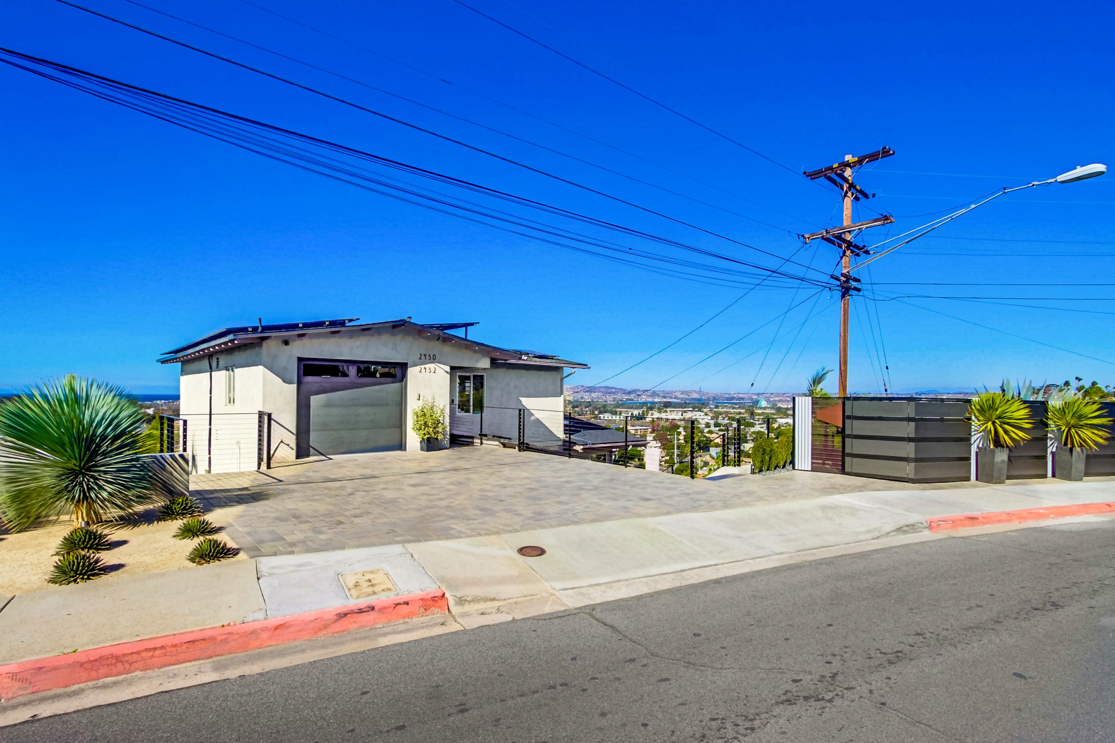 A modern house with a paved driveway is situated on a hilltop, overlooking the city under a clear blue sky.