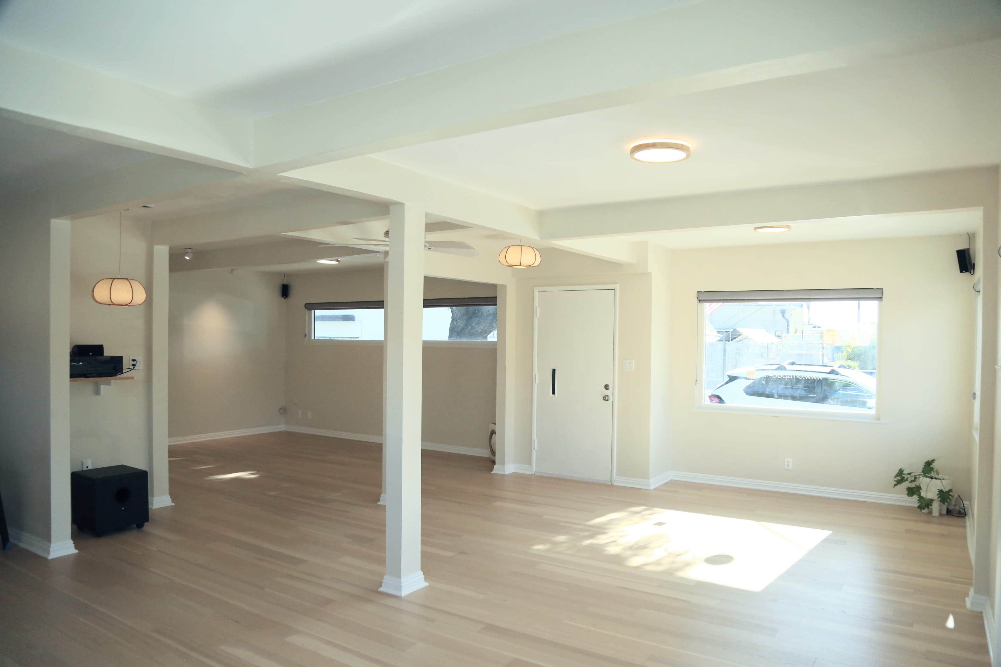The image shows a spacious, empty room with light hardwood flooring, large windows, and modern ceiling lights.