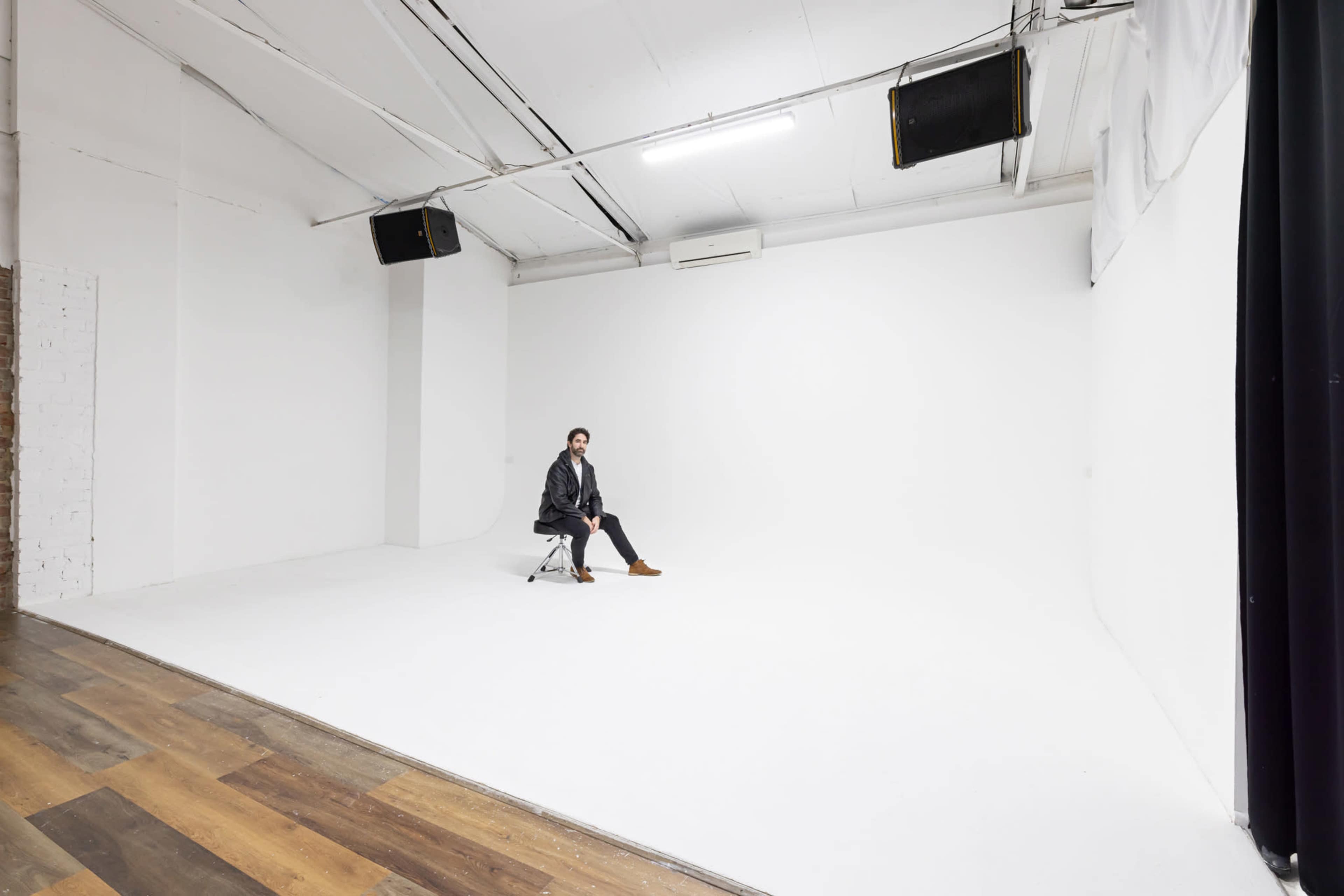 A person sits on a chair in an empty, well-lit studio with white walls and a wooden floor.