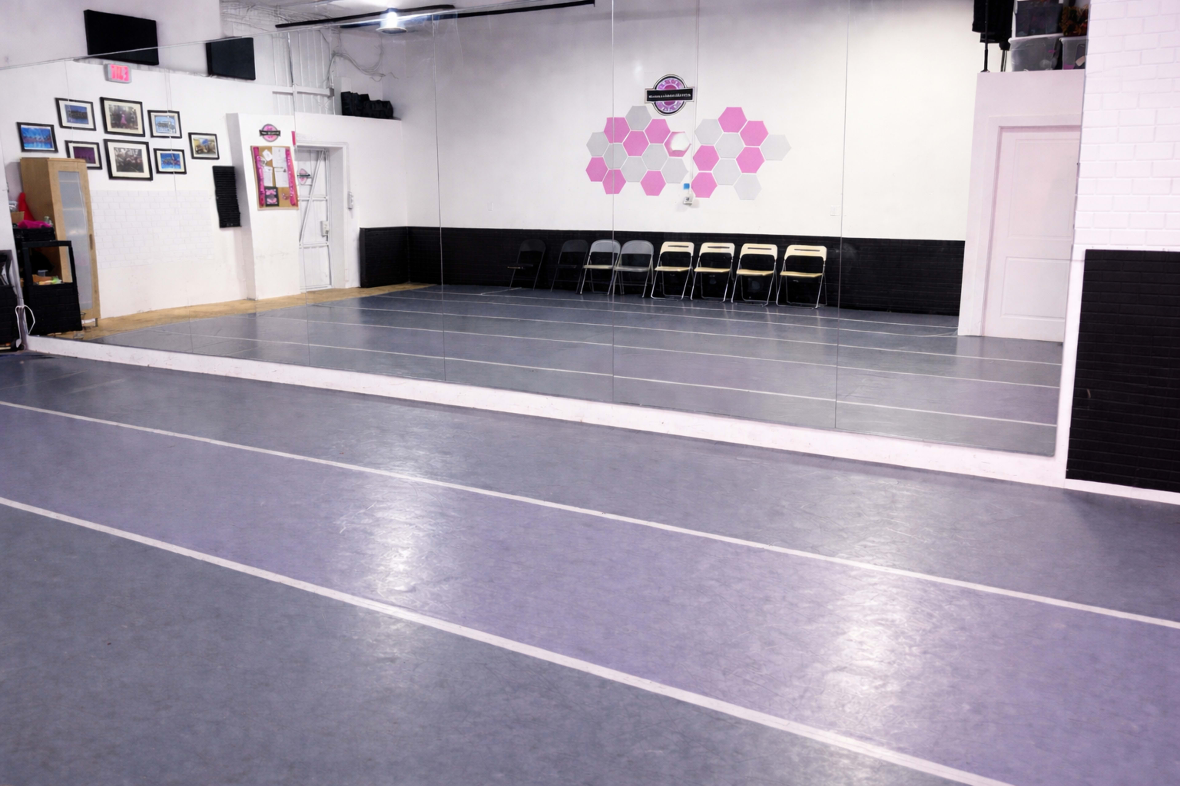 The image shows a dance studio with a mirrored wall, gray flooring, and a row of black folding chairs along the side.