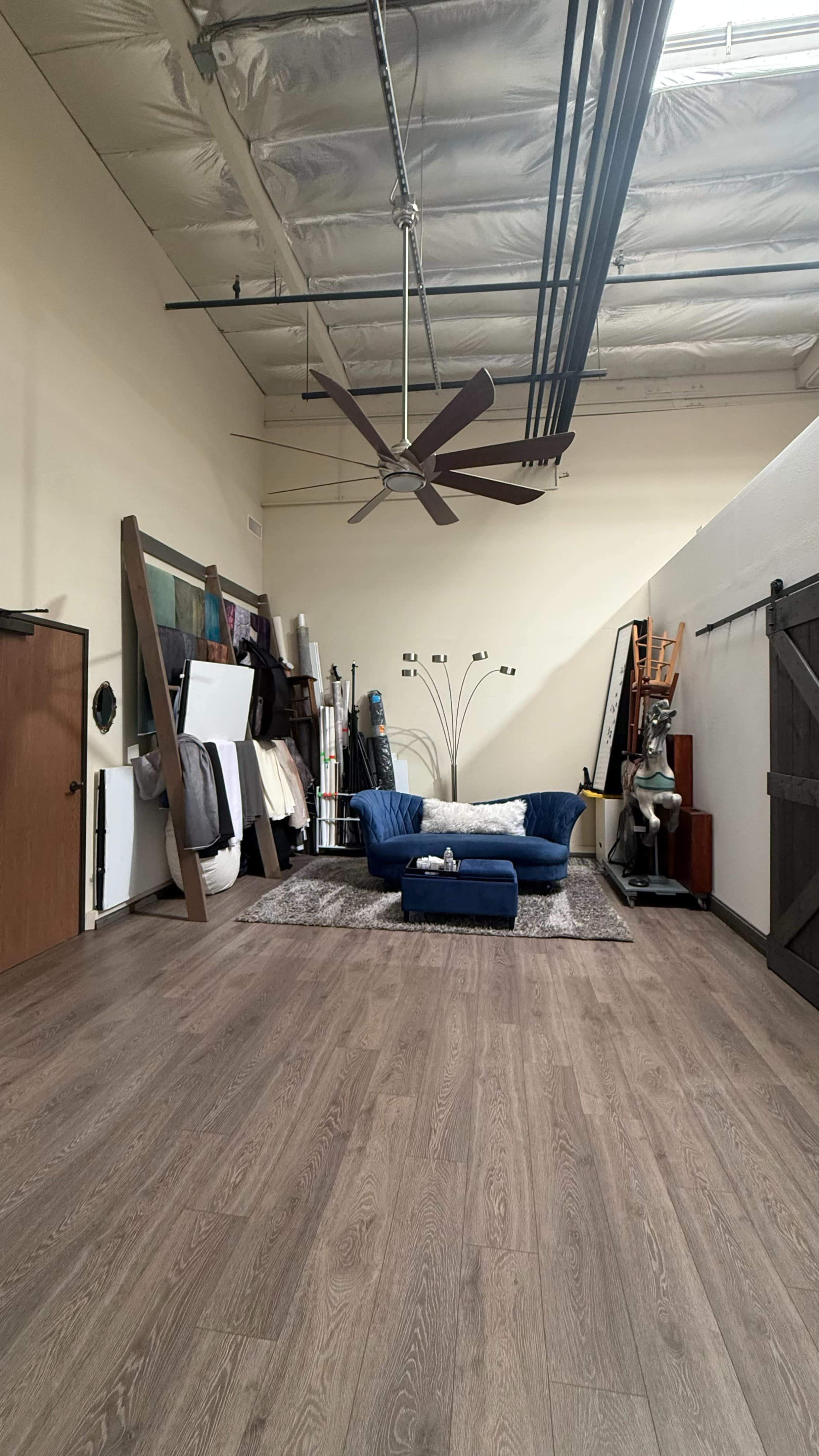 The image shows a spacious room with wooden flooring, featuring a blue couch, a large ceiling fan, and various items arranged around the area.