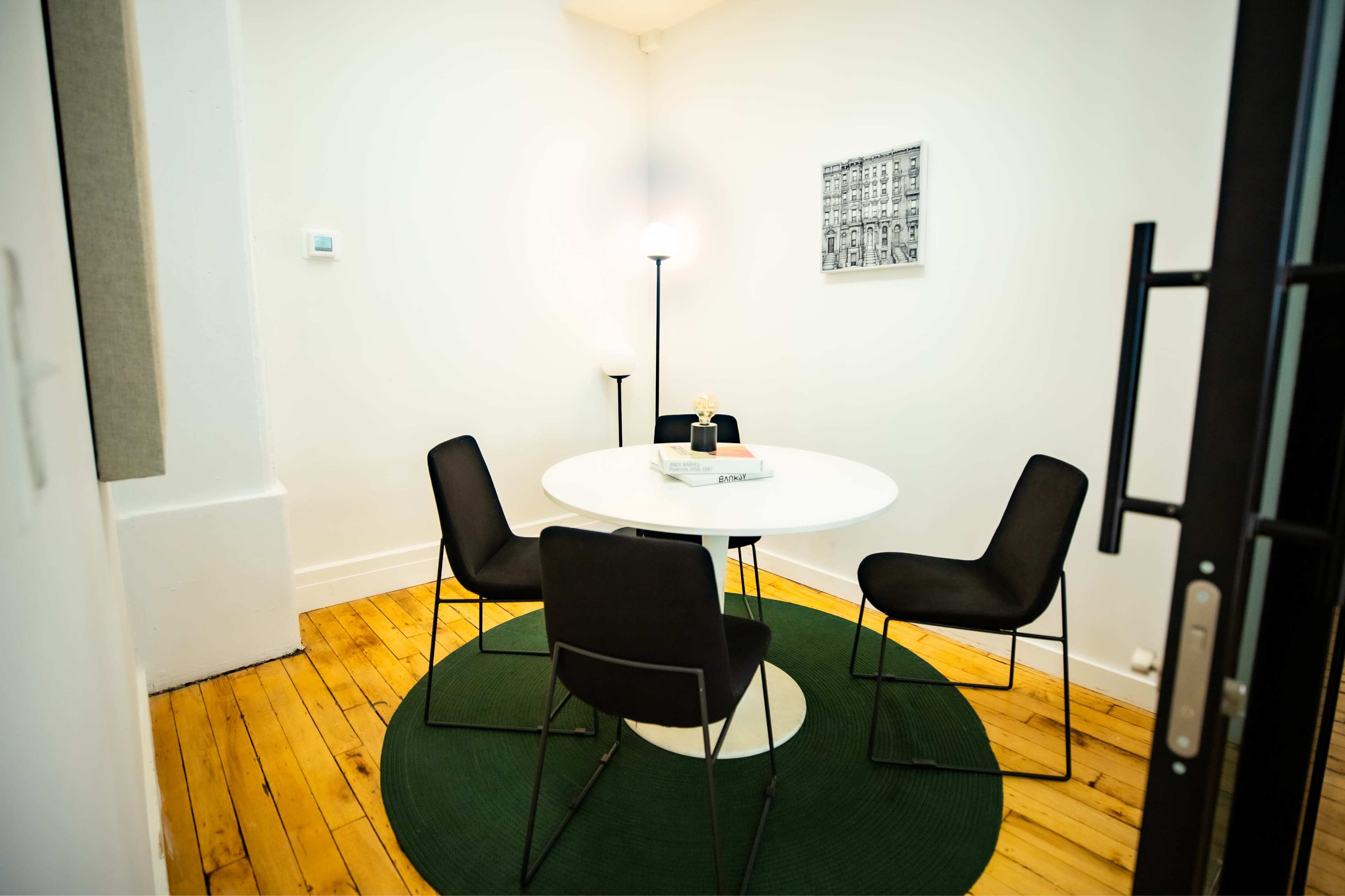 A small meeting room features a round white table surrounded by four black chairs on a green circular rug, with a lamp and framed photograph on the walls.