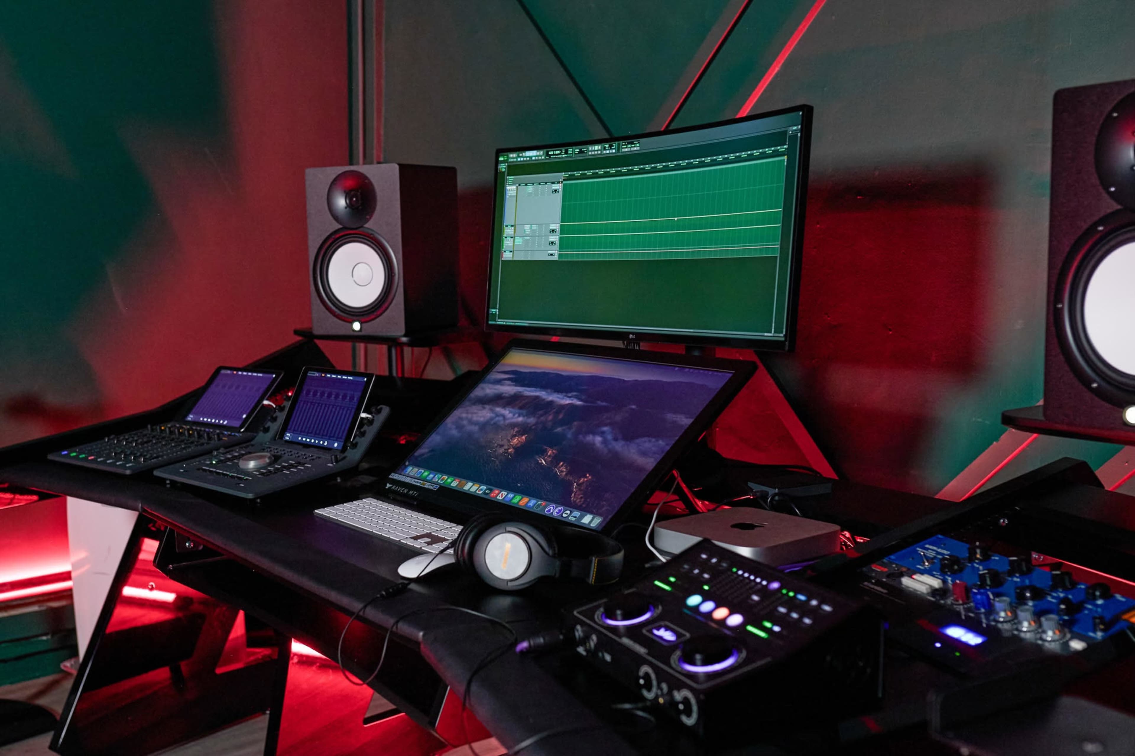 A music production setup featuring multiple monitors, audio interfaces, speakers, and a laptop arranged on a black desk with colored lighting in the background.