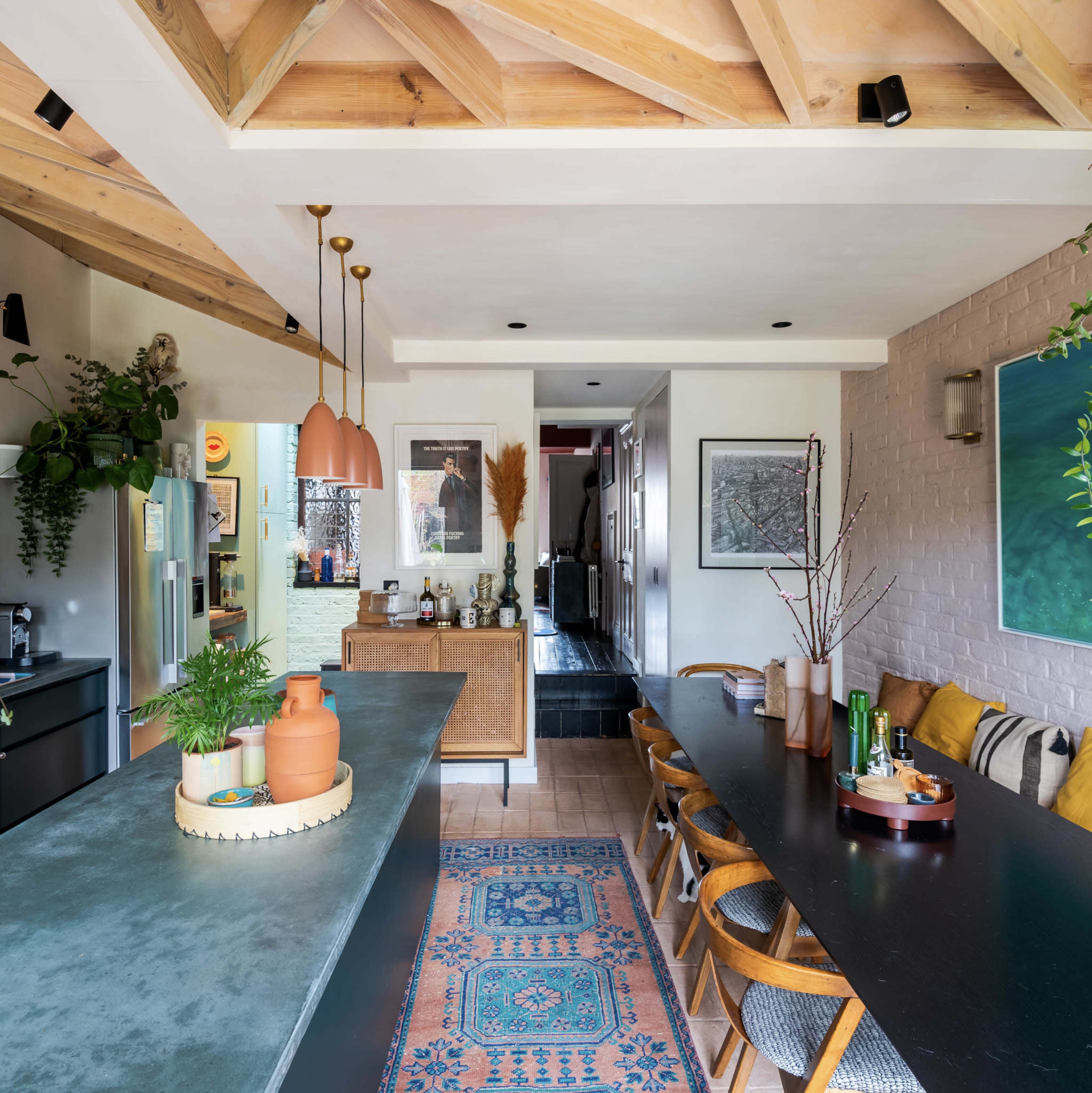 A modern kitchen and dining area featuring a long black dining table, wooden beams in the ceiling, and various plants and decor items throughout.