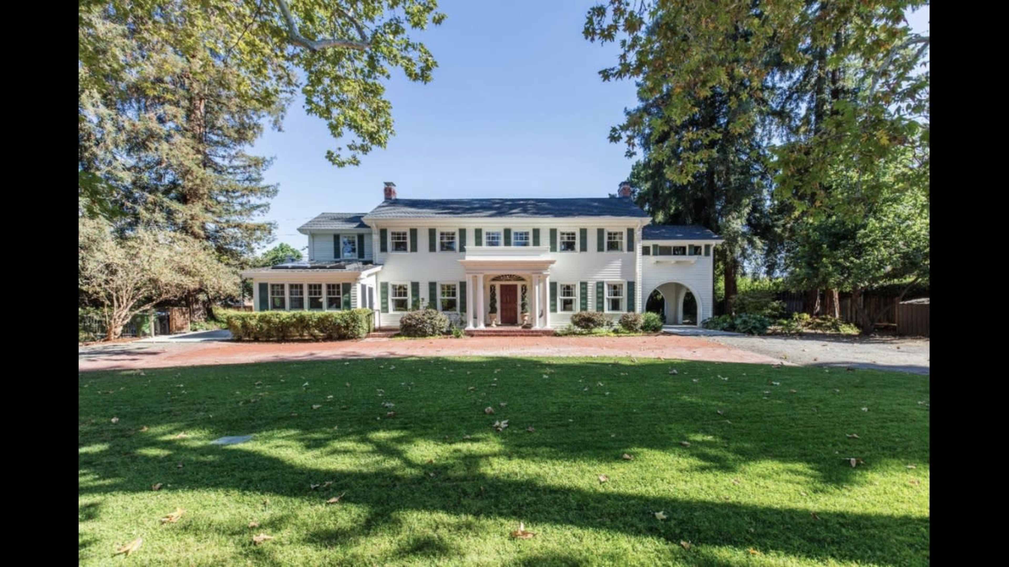 A large, two-story white house with a centered entrance and a green lawn is surrounded by trees and a gravel driveway.