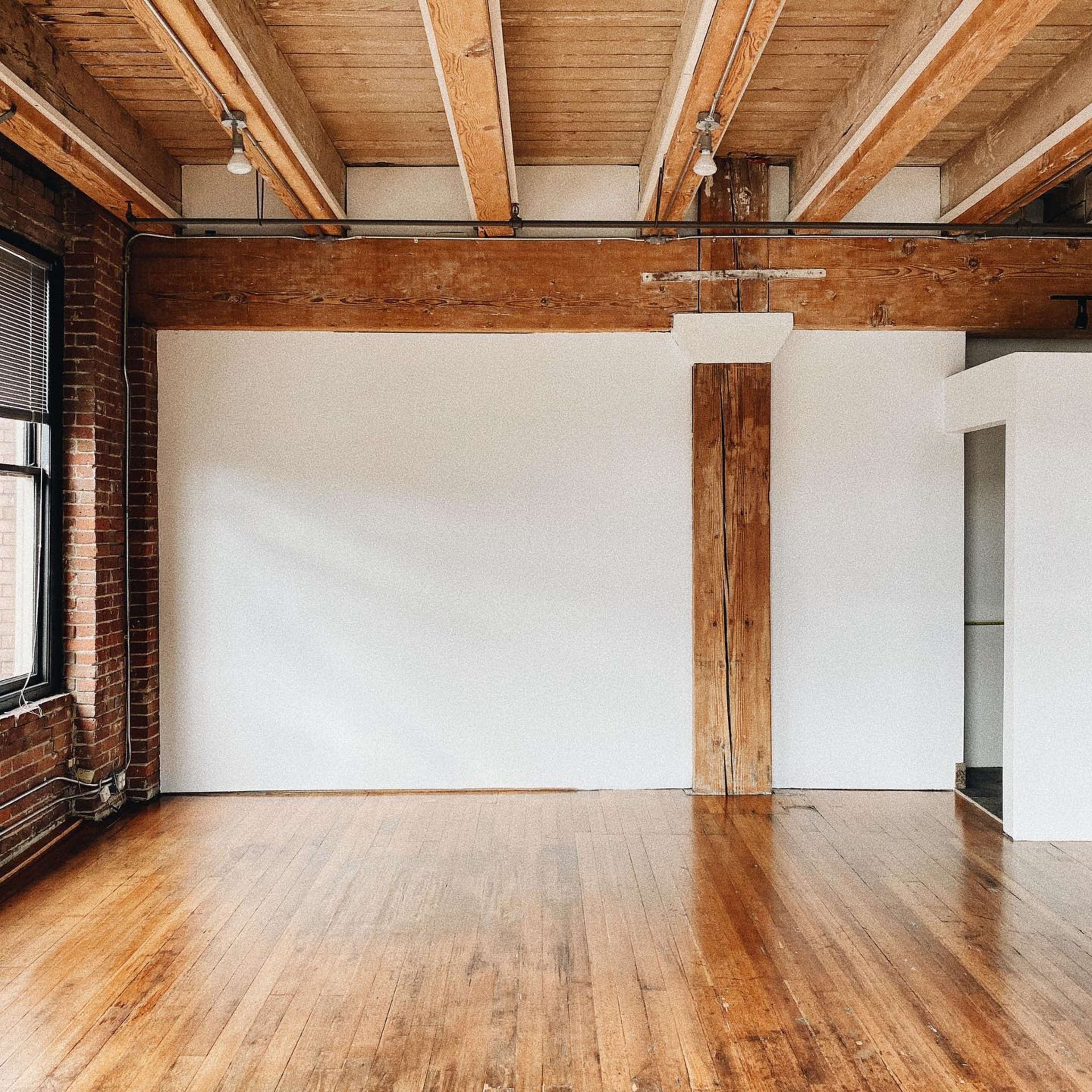 The image shows an empty room with exposed wooden beams, brick walls, and a polished wooden floor.
