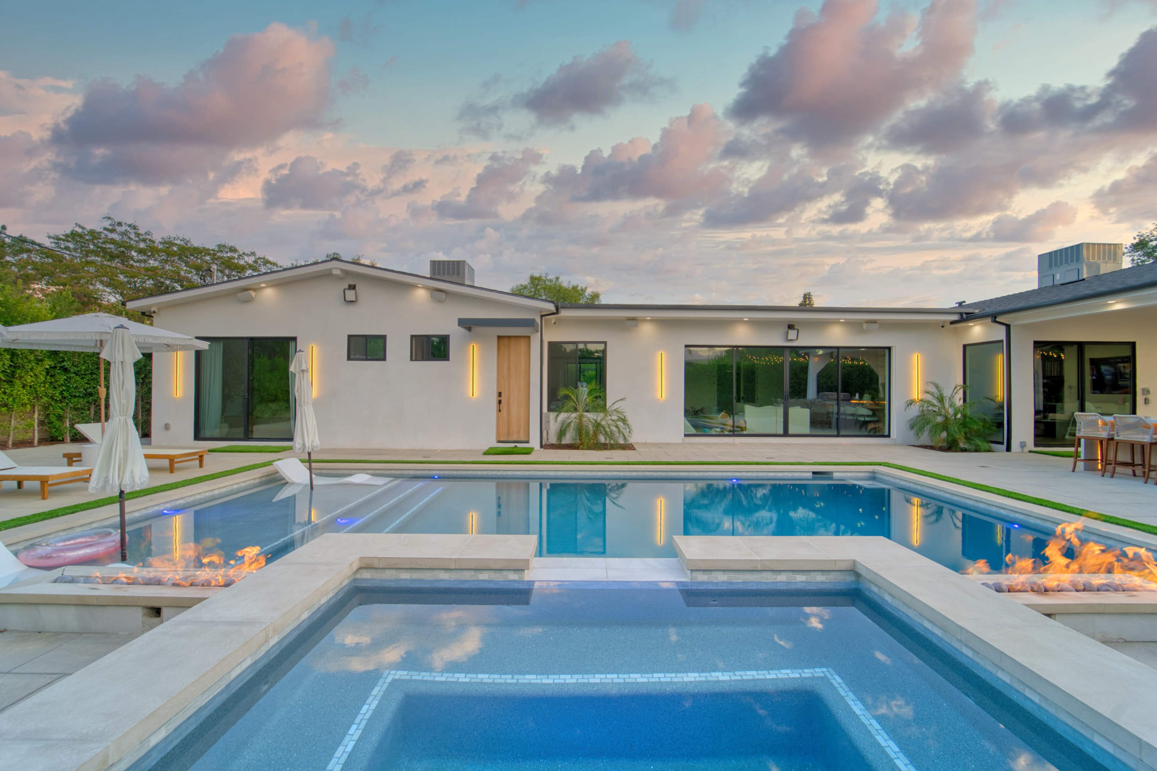 A modern house with large windows surrounds a rectangular swimming pool, featuring a fire pit and lounge chairs beside it.