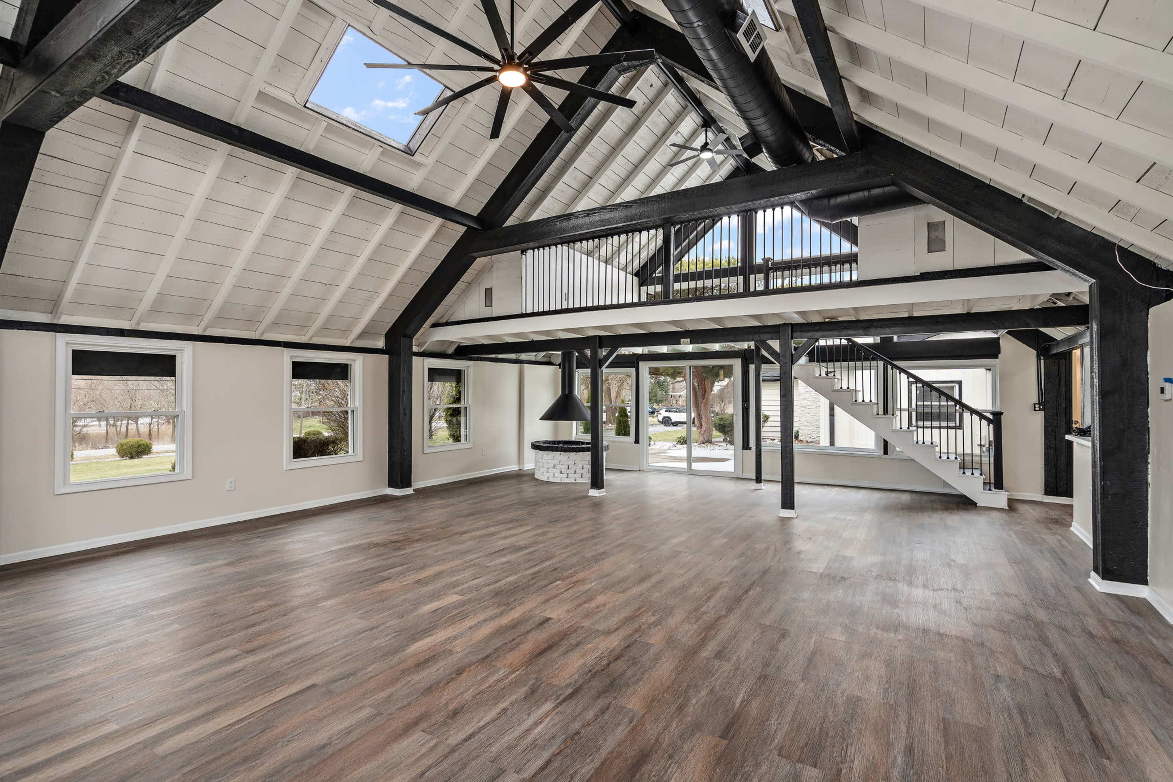 The image shows a spacious, open interior with a high ceiling supported by black beams, large windows, and a staircase in the background.