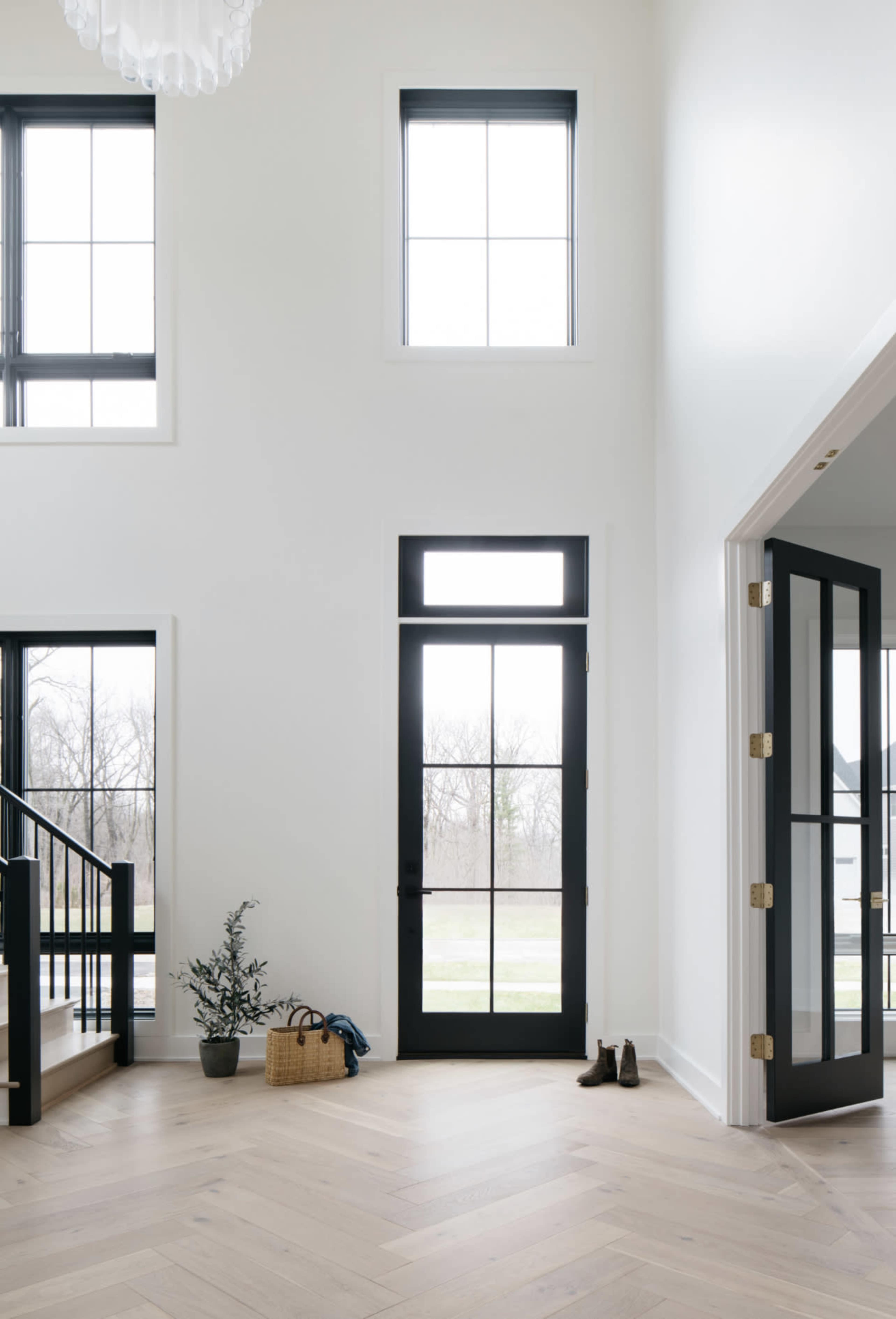 The image shows a spacious foyer with large windows, a black front door, a staircase, and minimal decor including a small plant and a basket.