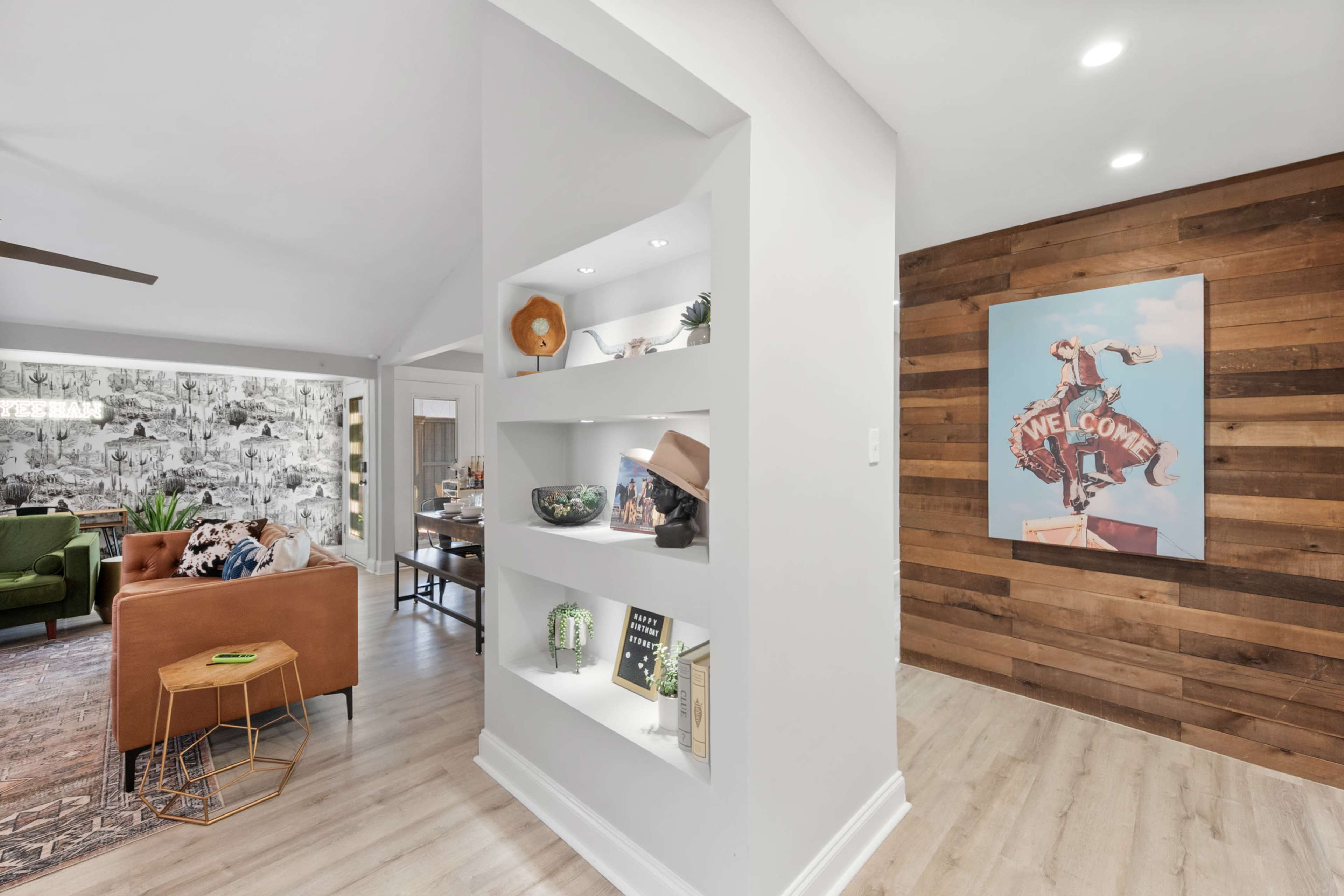 The image shows a modern living space with a sectional sofa, decorative shelves, and a wooden accent wall featuring a retro-style artwork.