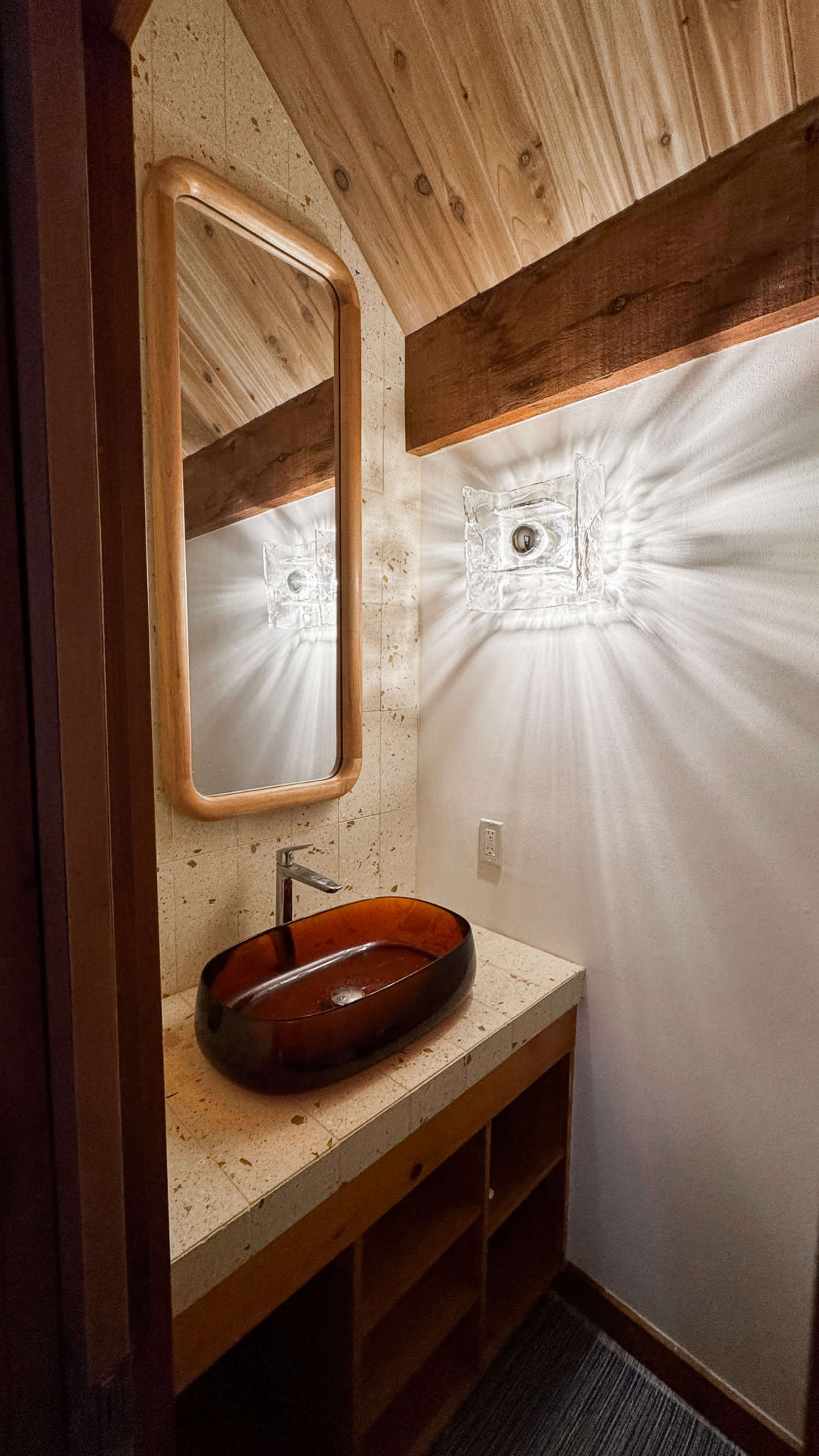 The image shows a bathroom featuring a wooden vessel sink on a stone countertop, a large mirror, and textured wall lighting creating a radiant effect.